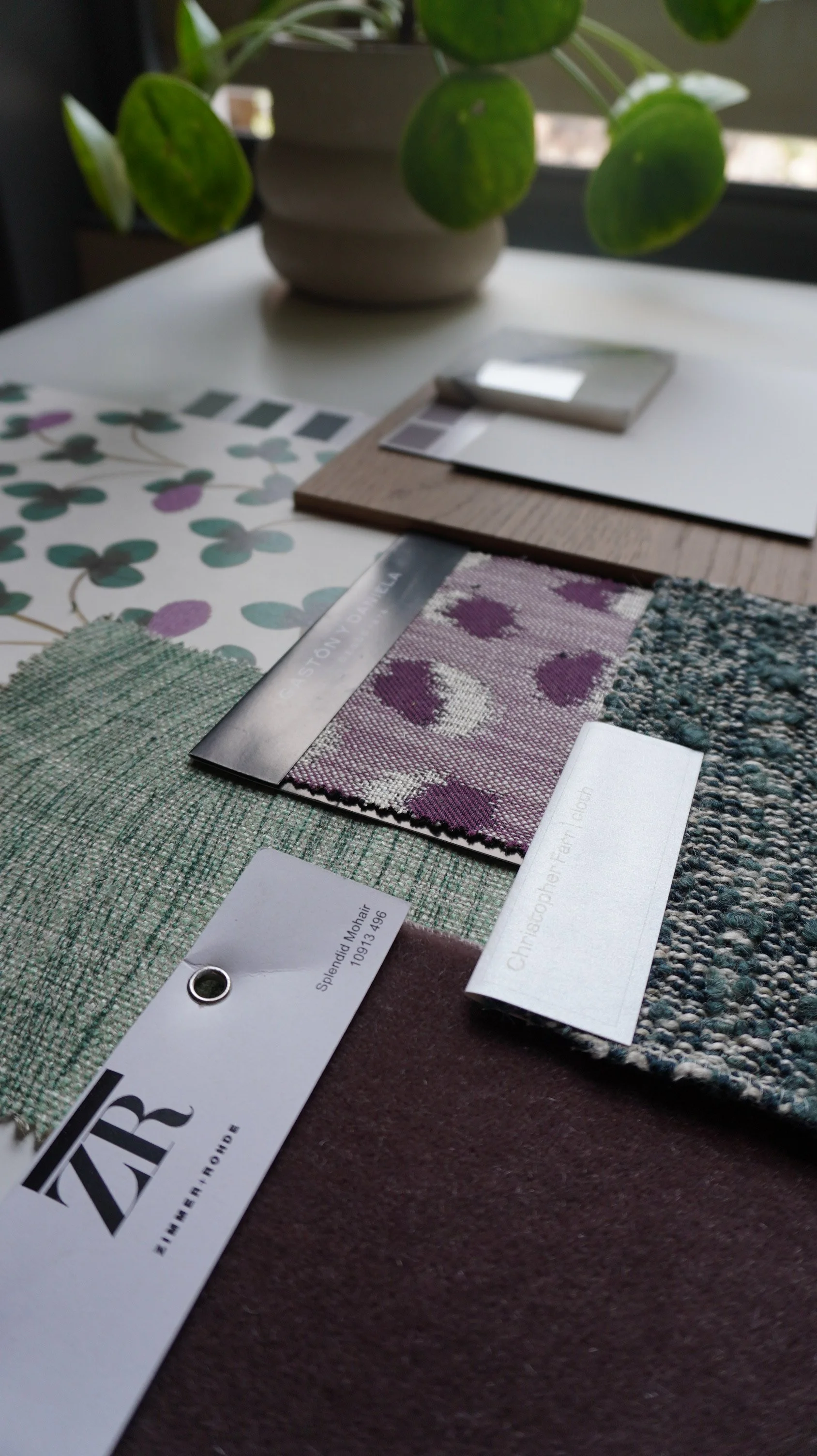 Close-up of fabric samples with various textures and patterns, arranged on a table, with a plant in the background.