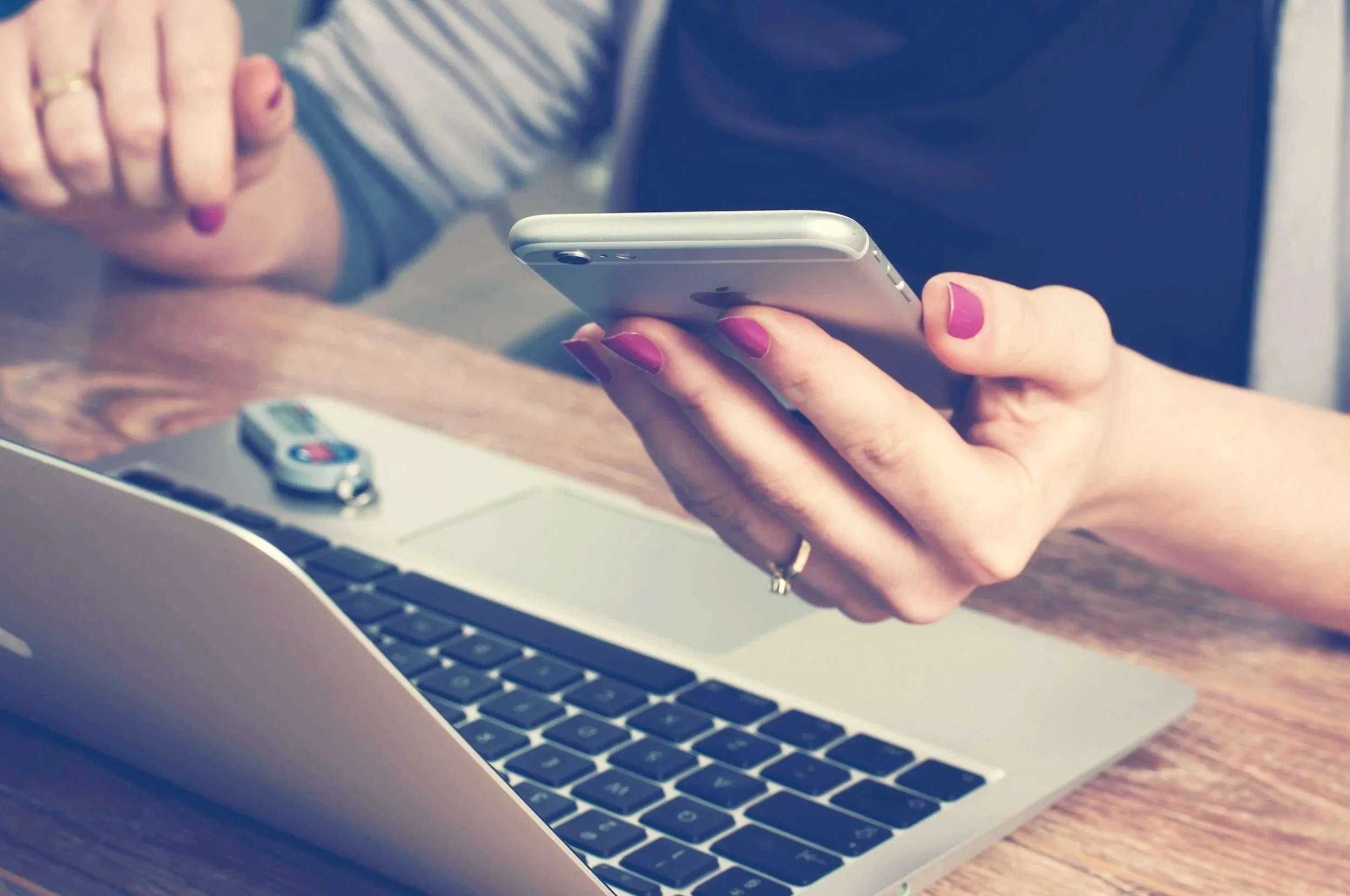 Close-up of a person's hands holding a smartphone above a laptop keyboard, with a watch and a remote control on the table.