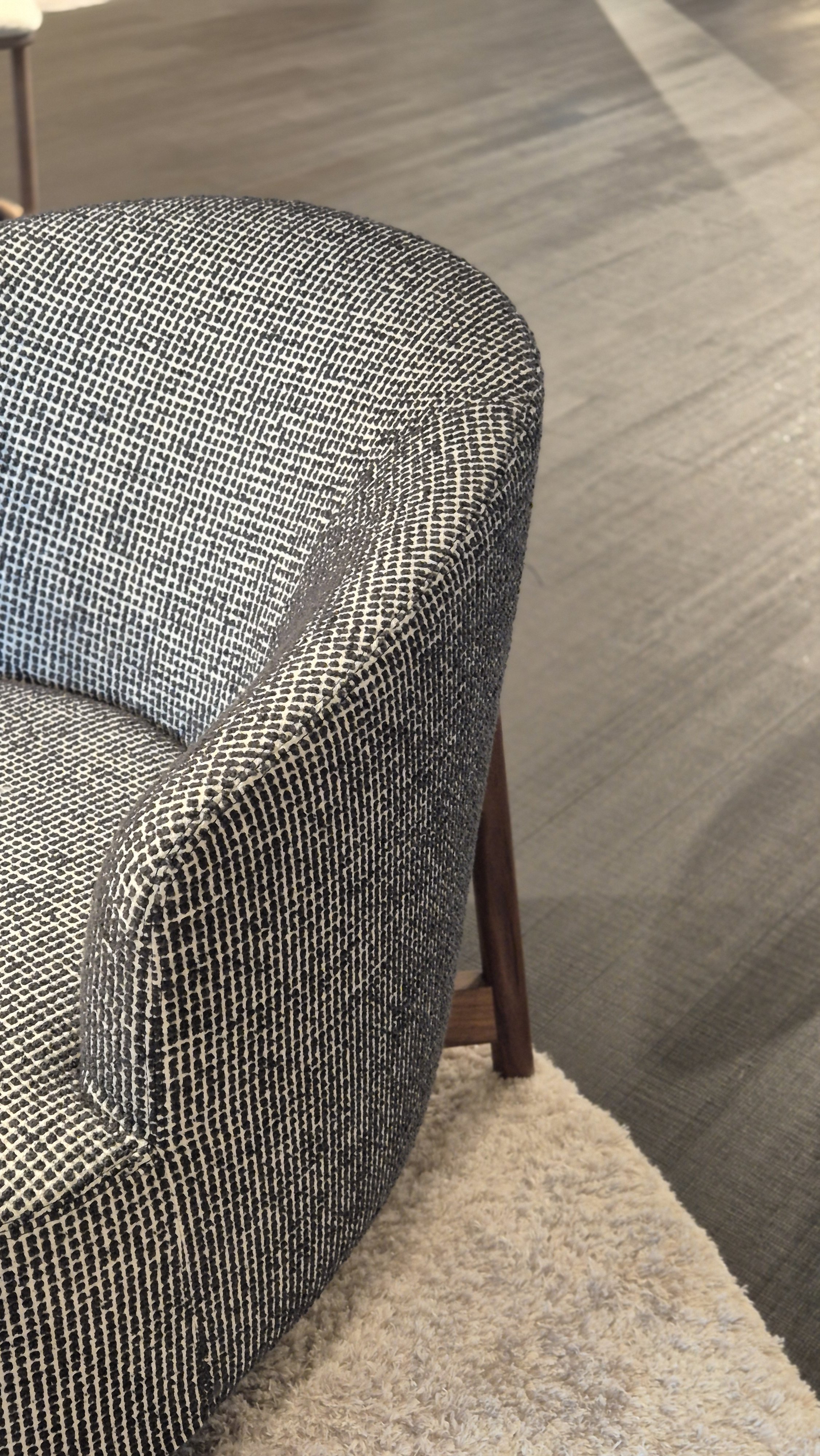 Close-up view of a textured, patterned upholstered chair with wooden legs, placed on a plush, cream-colored rug on a dark wooden floor.
