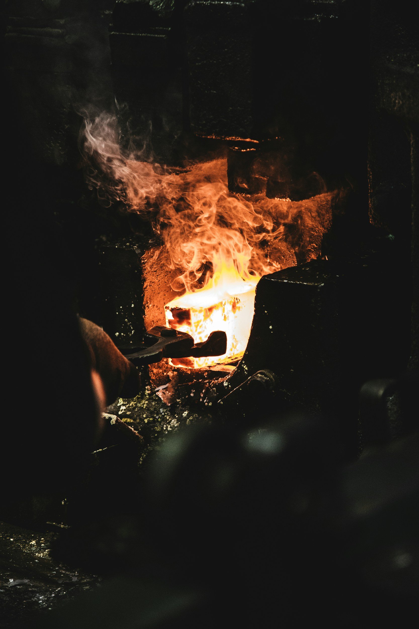 A person holding black tongs in front of a forge fire with bright orange flames and glowing hot metal inside a blacksmithing forge.