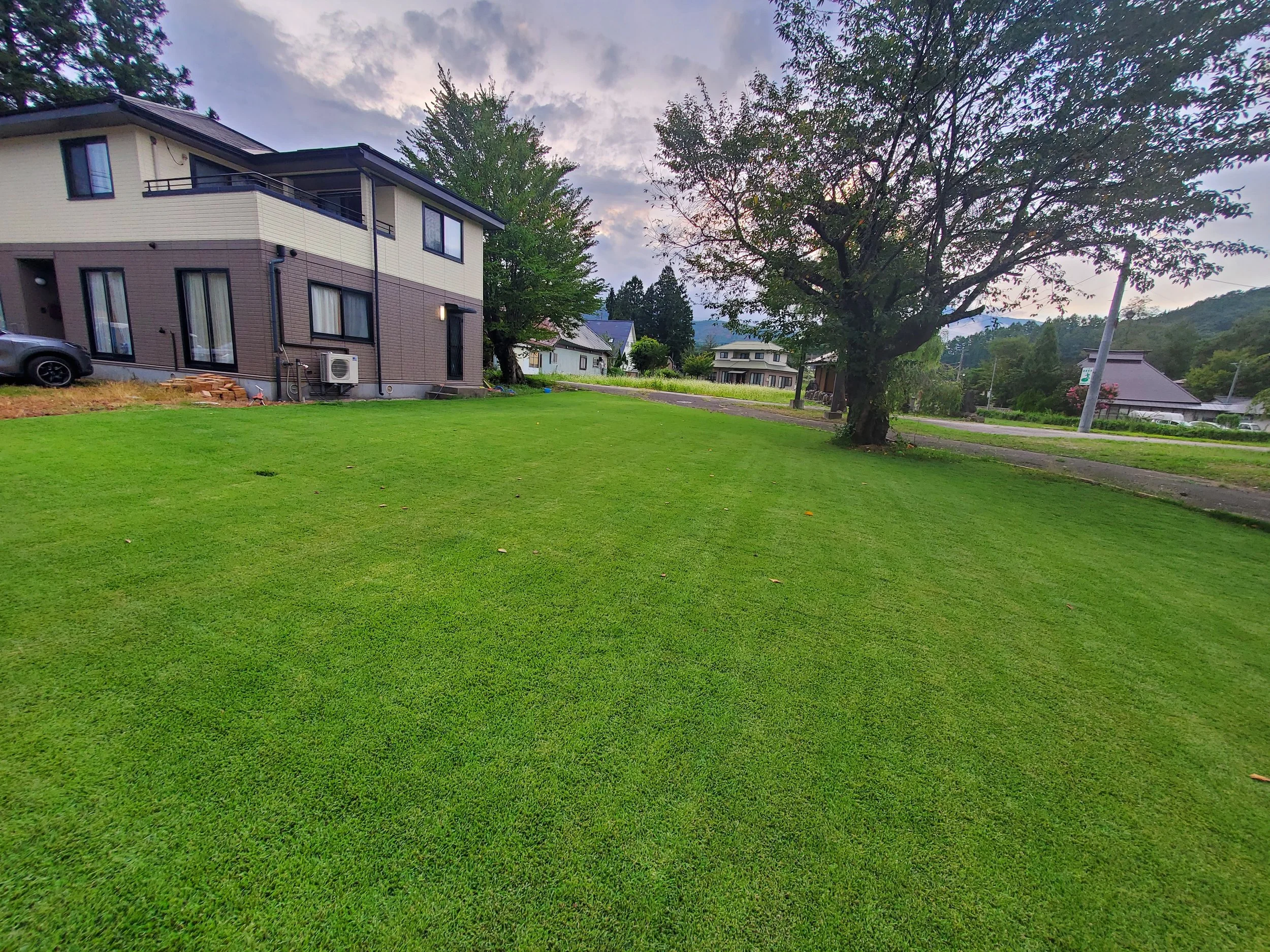 A suburban scene with a modern two-story house on the left, featuring a brown and beige exterior, a car parked in front, and an air conditioning unit attached to the wall. The yard has a well-maintained green lawn and a large leafy tree on the right. In the background, there are other houses, trees, and hills under a partly cloudy sky.