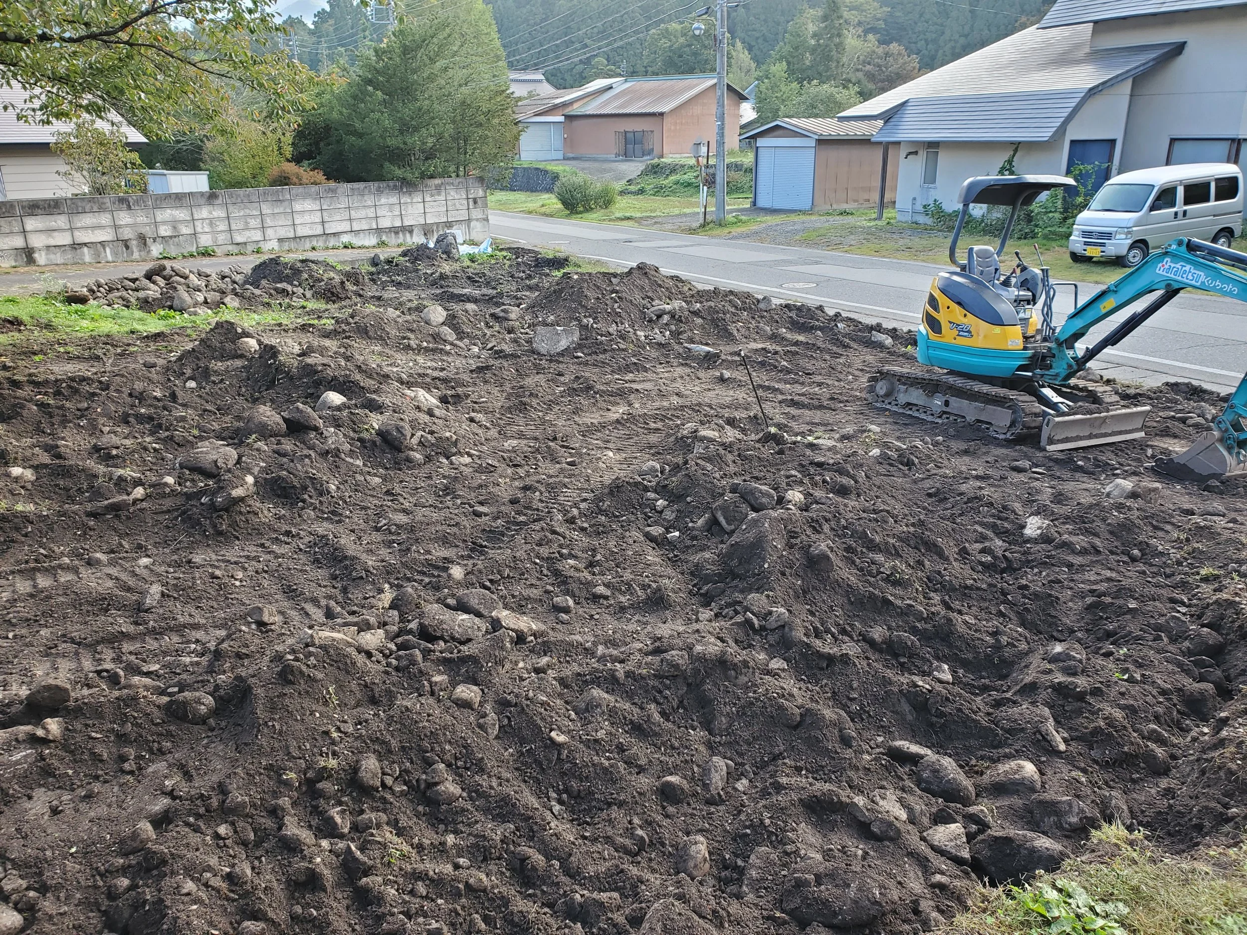 A construction site with uneven dirt and rocks, and a small blue and yellow excavator parked on the right. In the background, there are residential houses, trees, a concrete block wall, and power lines.