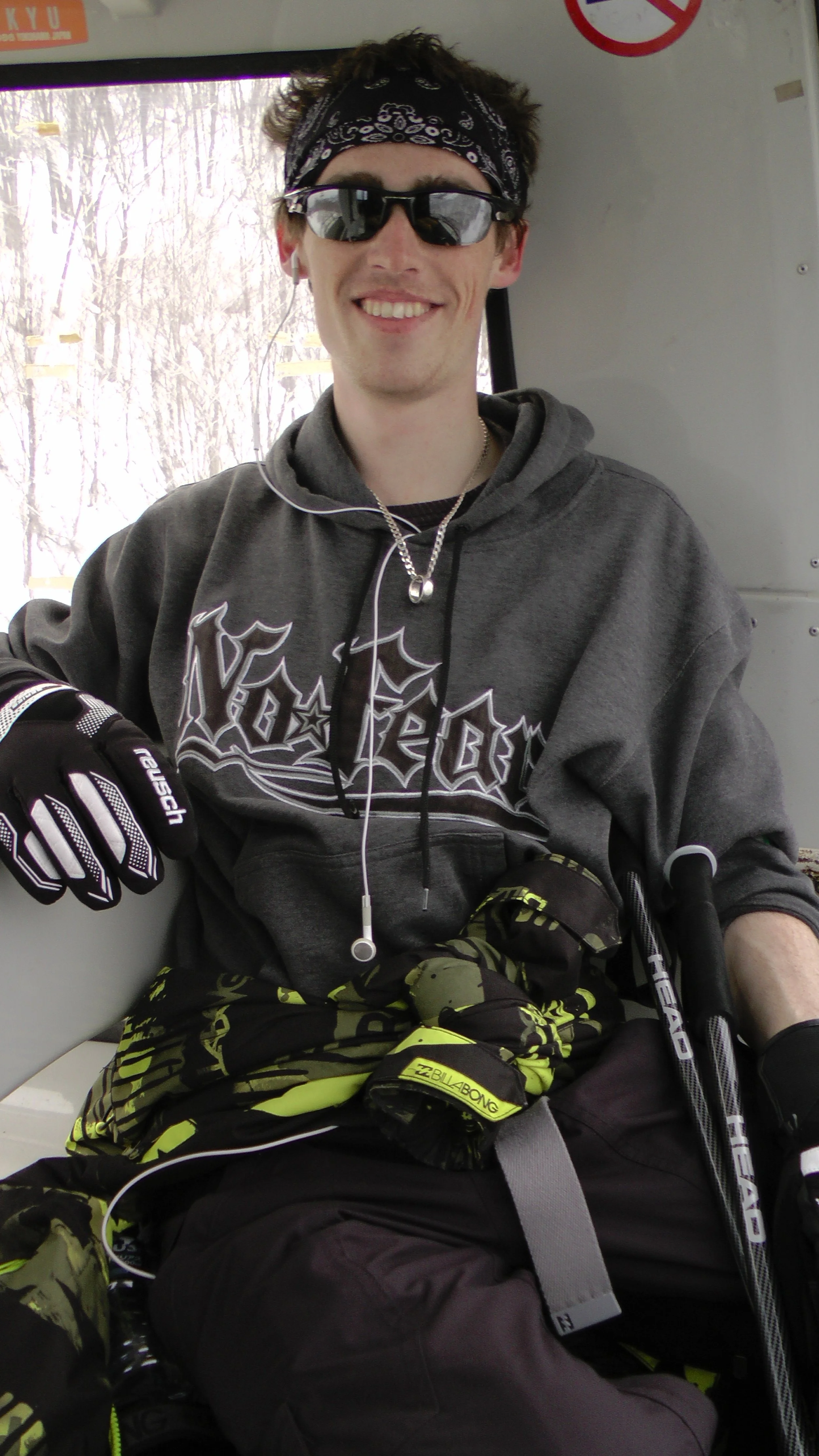 A young man with curly hair wearing sunglasses, a black bandana, and sweatshirts with a graphic design, sitting inside a vehicle with a snowy scene outside the window.