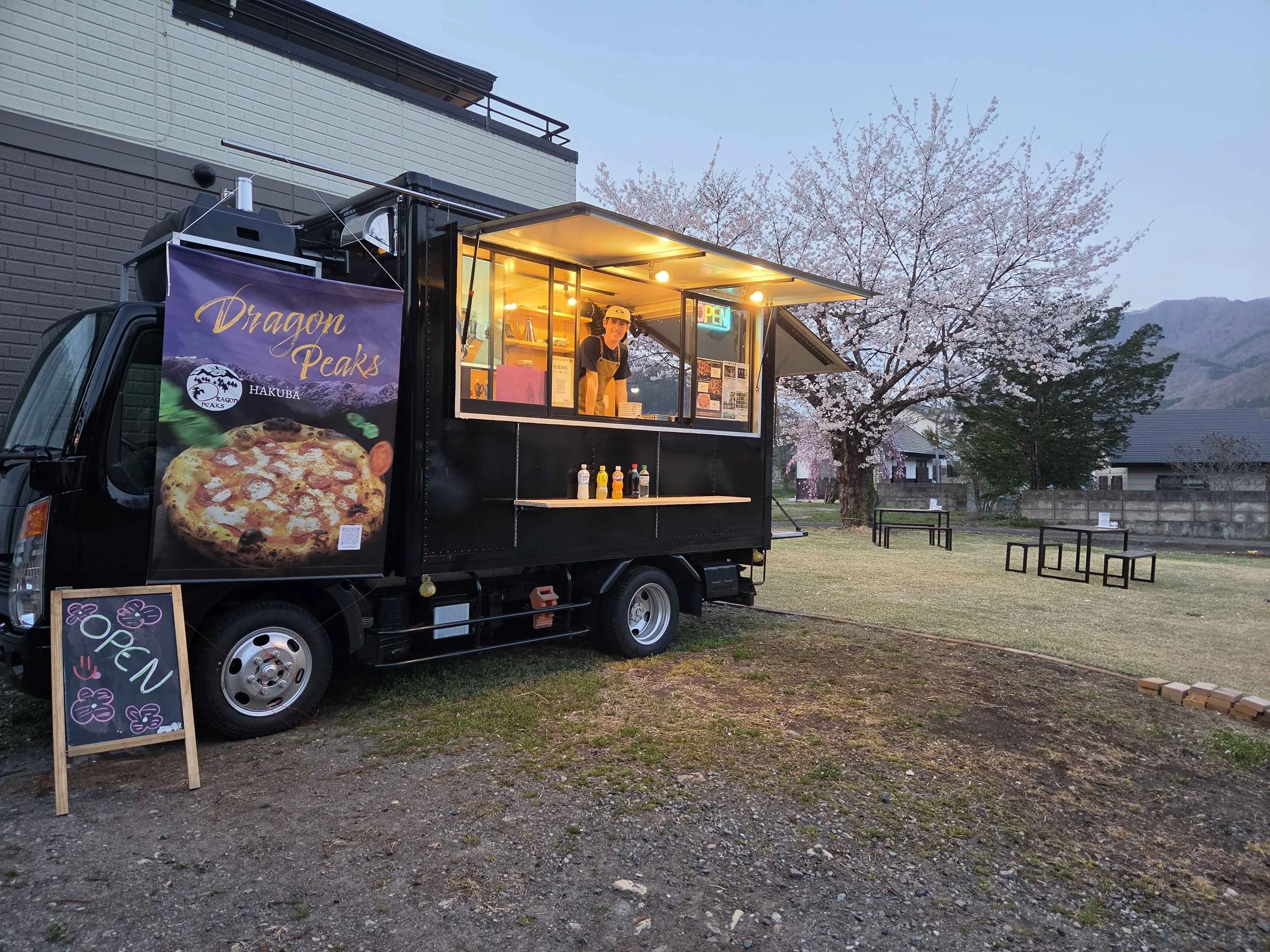 A black food truck with a sign for pizza and a banner that reads 'Dragon Peaks' parked outdoors near cherry blossom trees. The truck has a small window with a person inside and condiments on a shelf.