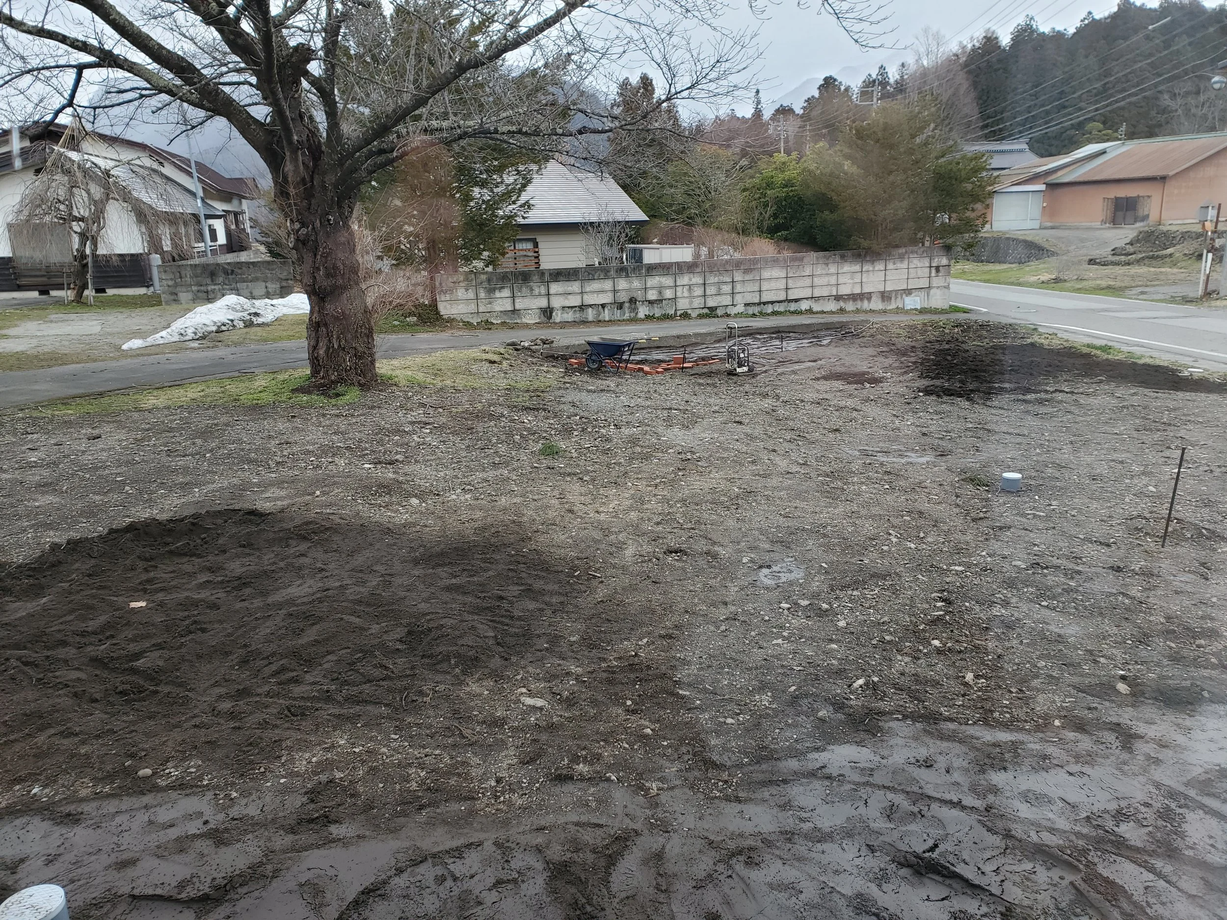 A construction site with dug-up soil in the foreground, a wheelbarrow, and construction tools. In the background, there are houses, trees, and mountains.