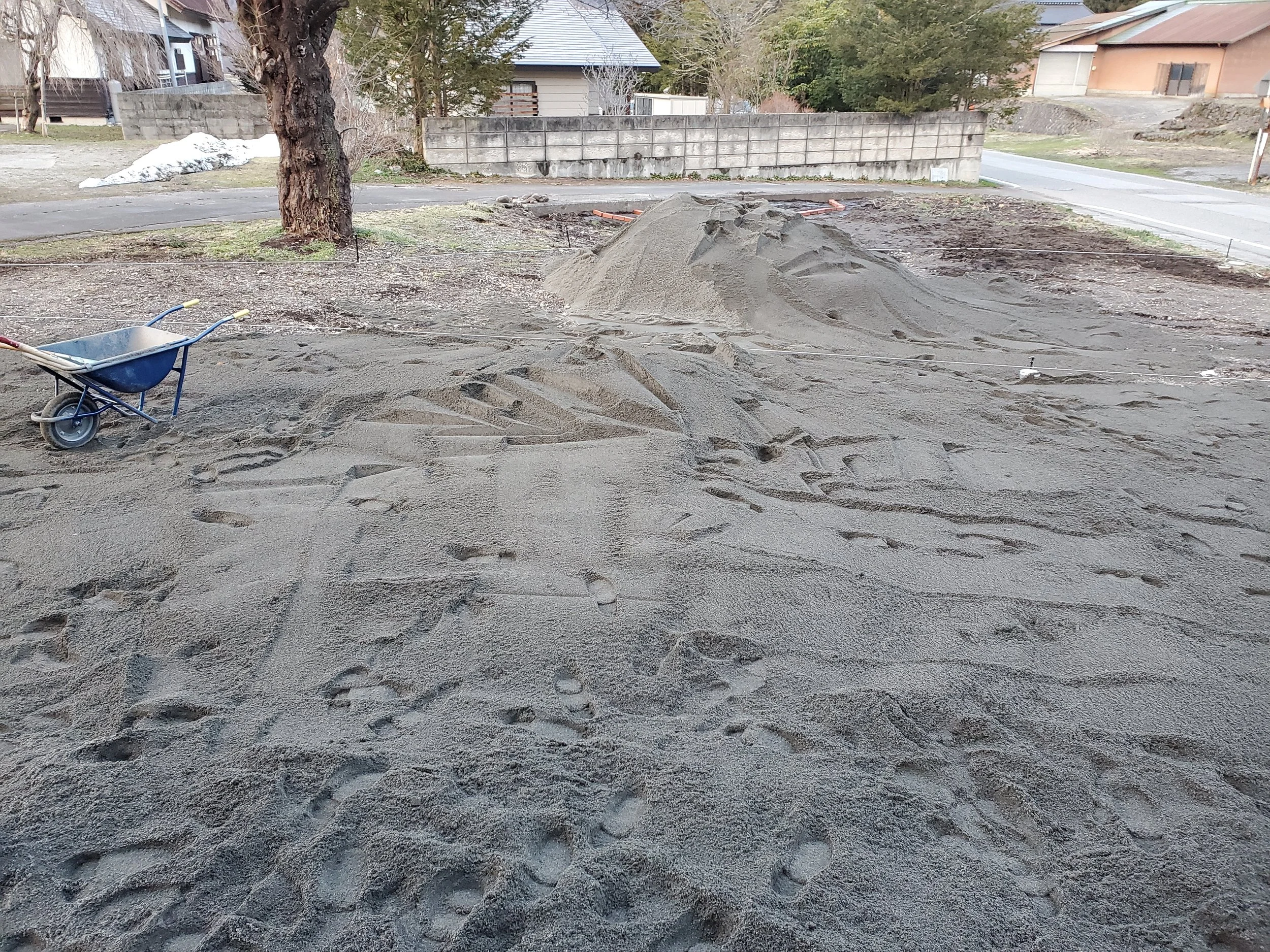 Construction site with a pile of sand, a wheelbarrow, some disturbed soil, a tree, and a sidewalk with residential houses in the background.