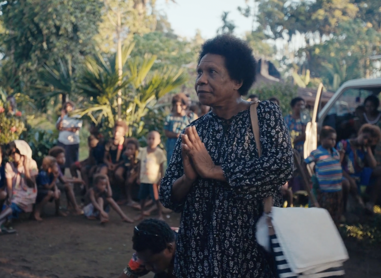 An elder woman, Lydia Gah of the Nakanai community stands with her hands pressed together in a praying gesture, outdoors among a group of children and other people in a Baikakea village with ancestral trees in the background.