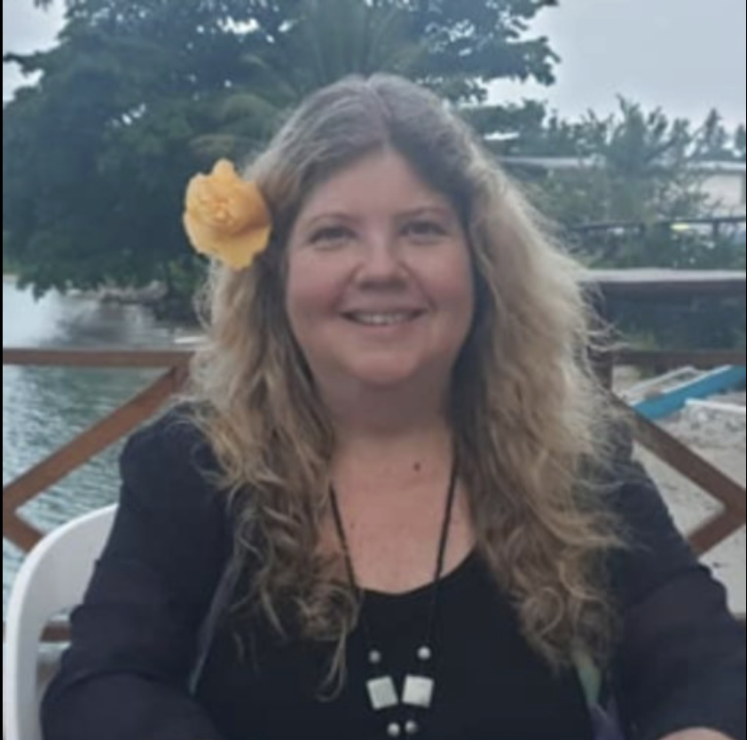 A woman with long curly hair and a yellow flower in her hair, smiling while sitting outdoors near water with trees in the background.