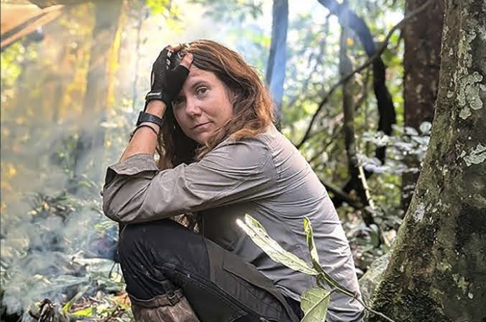 A woman sitting on the forest ground, resting against a tree, with a serious expression, wearing outdoor gear and gloves, surrounded by trees and foliage.