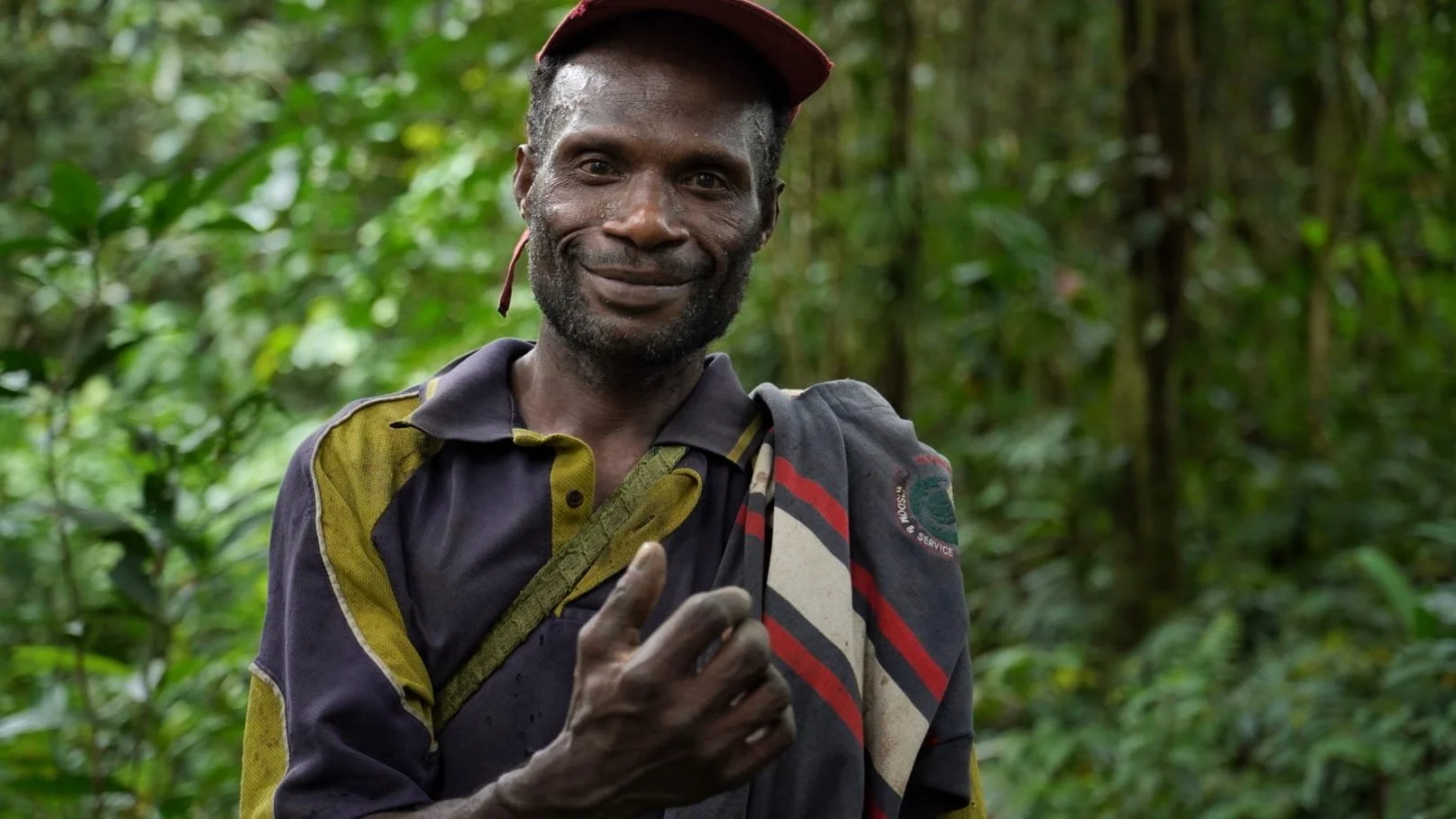 A smiling man in a dark shirt with yellow and red accents, wearing a hat, standing in the lush green Lamo Auru forest.