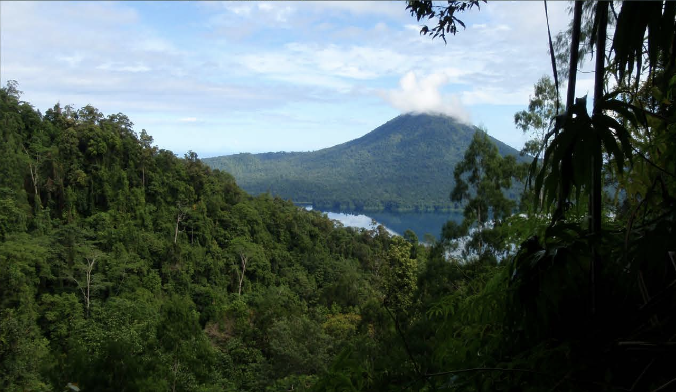 Lamo Auru Caldera, a lush green forest leading to a lake with a mountain in the background, topped with clouds.