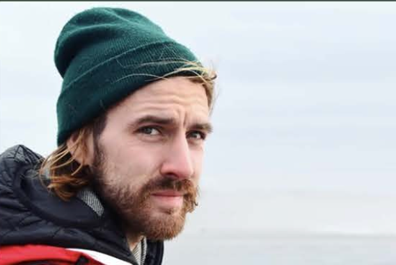 Close-up of a man wearing a green beanie and black jacket, outdoors on a cloudy day.