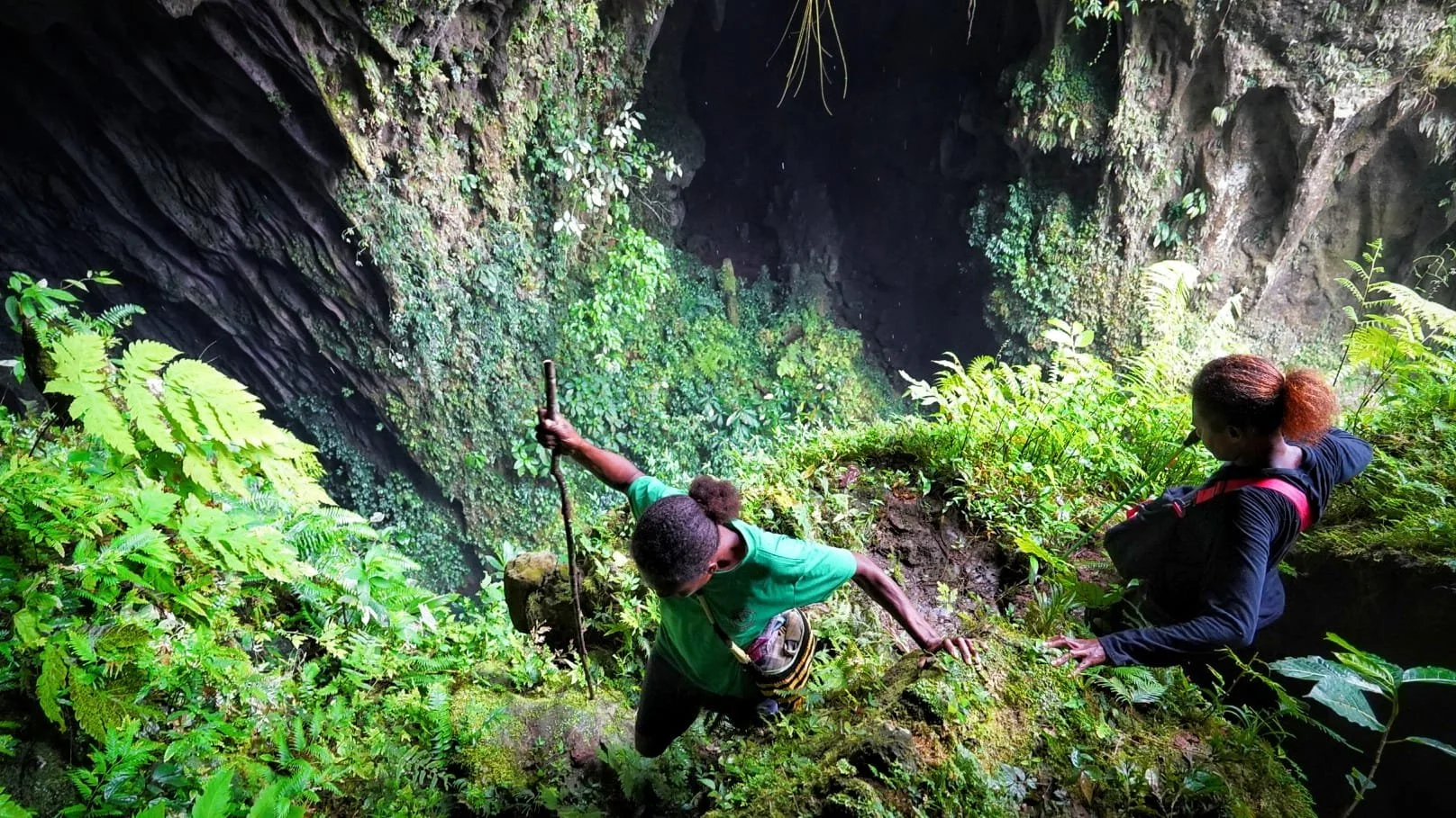 Two women hold onto a rope and climb out of a lush, green cave.