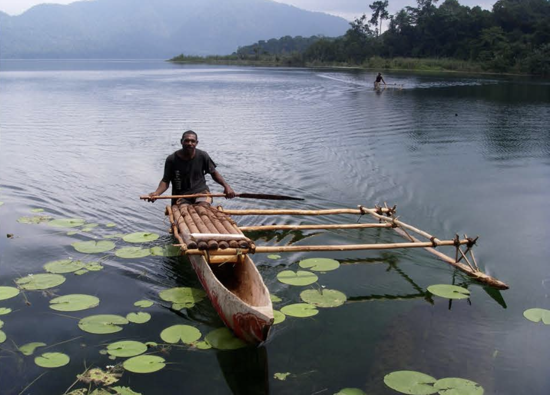 A man on a wooden raft on the blue lake, with another person on a canoe in the distance and mountains in the background.