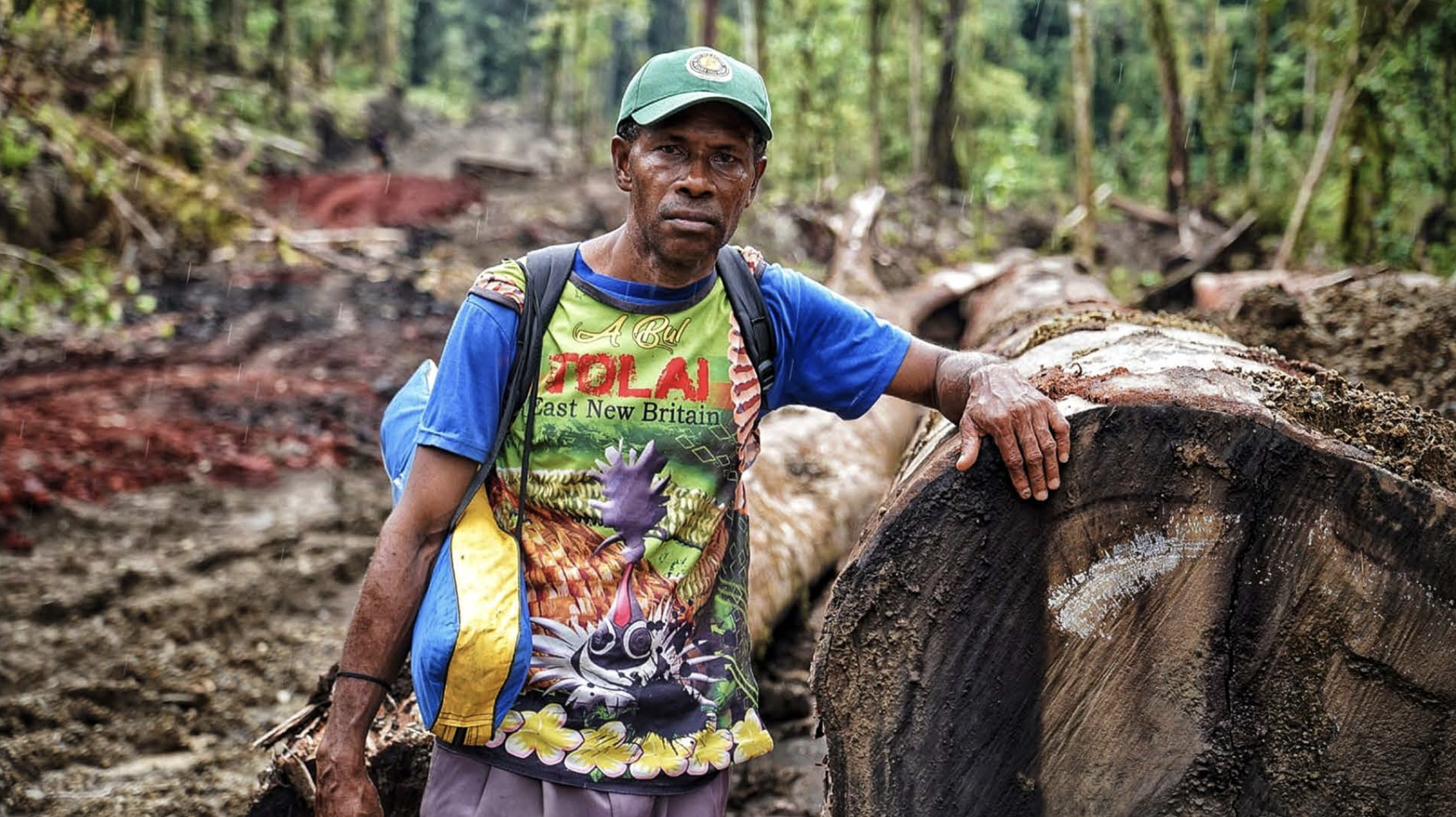 A Nakani Elder man with a blue shirt and colorful vest stands next to an illegally felled tree in a the Lamo Auru forest, with a concerned expression and a backpack.
