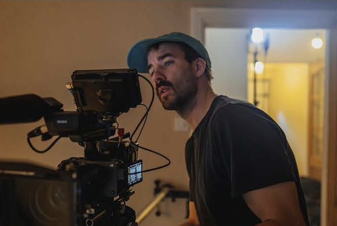 A man wearing a teal baseball cap and black t-shirt operating a professional movie camera indoors.