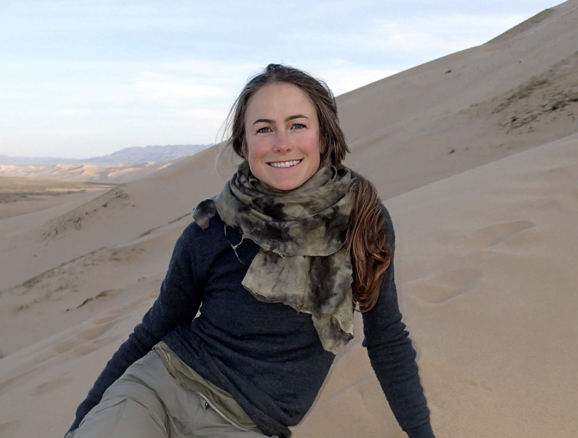 Smiling woman sitting on sand dunes in desert landscape with distant mountains under cloudy sky.