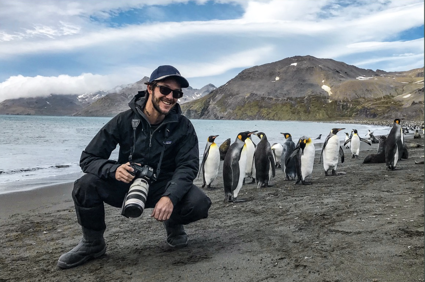 Ross Vernon McDonald crouches holding a longlens camera next to some penguins on a remote beach with mountains in the background