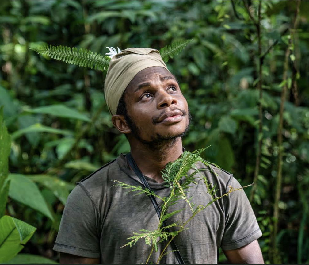 A man with a tan headband, bearded and wearing a dark shirt decorated with green ferns in the dense Lamo Auru jungle.