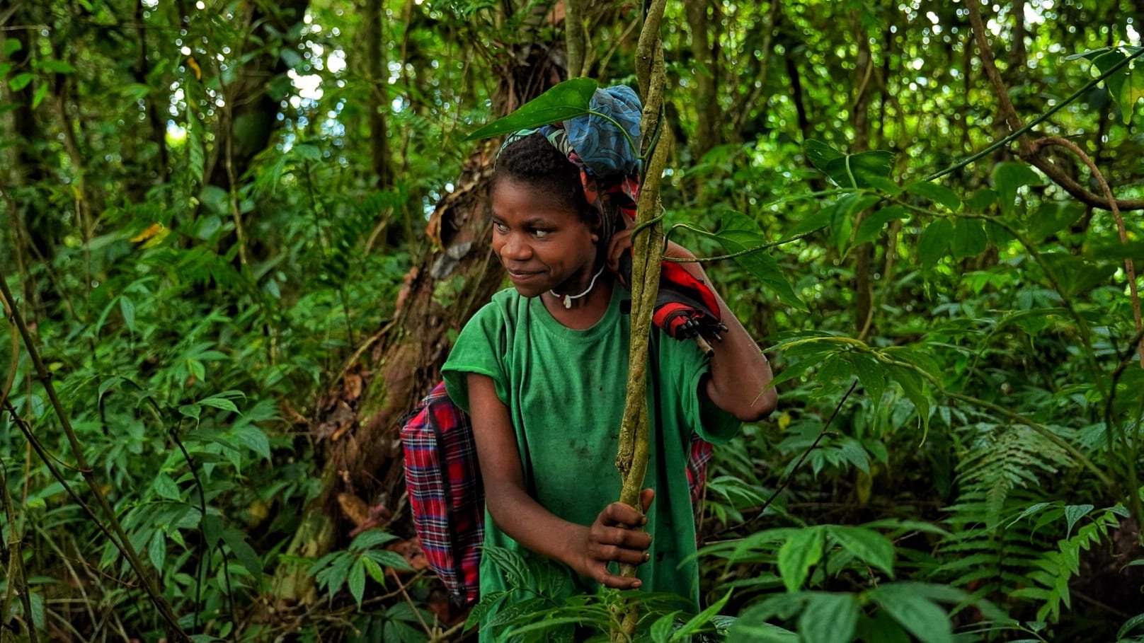 A young girl with short, styled hair, wearing a green t-shirt and a red plaid backpack, is in a dense green Lamo Auru jungle, holding a long stick and looking to her left with a smile.
