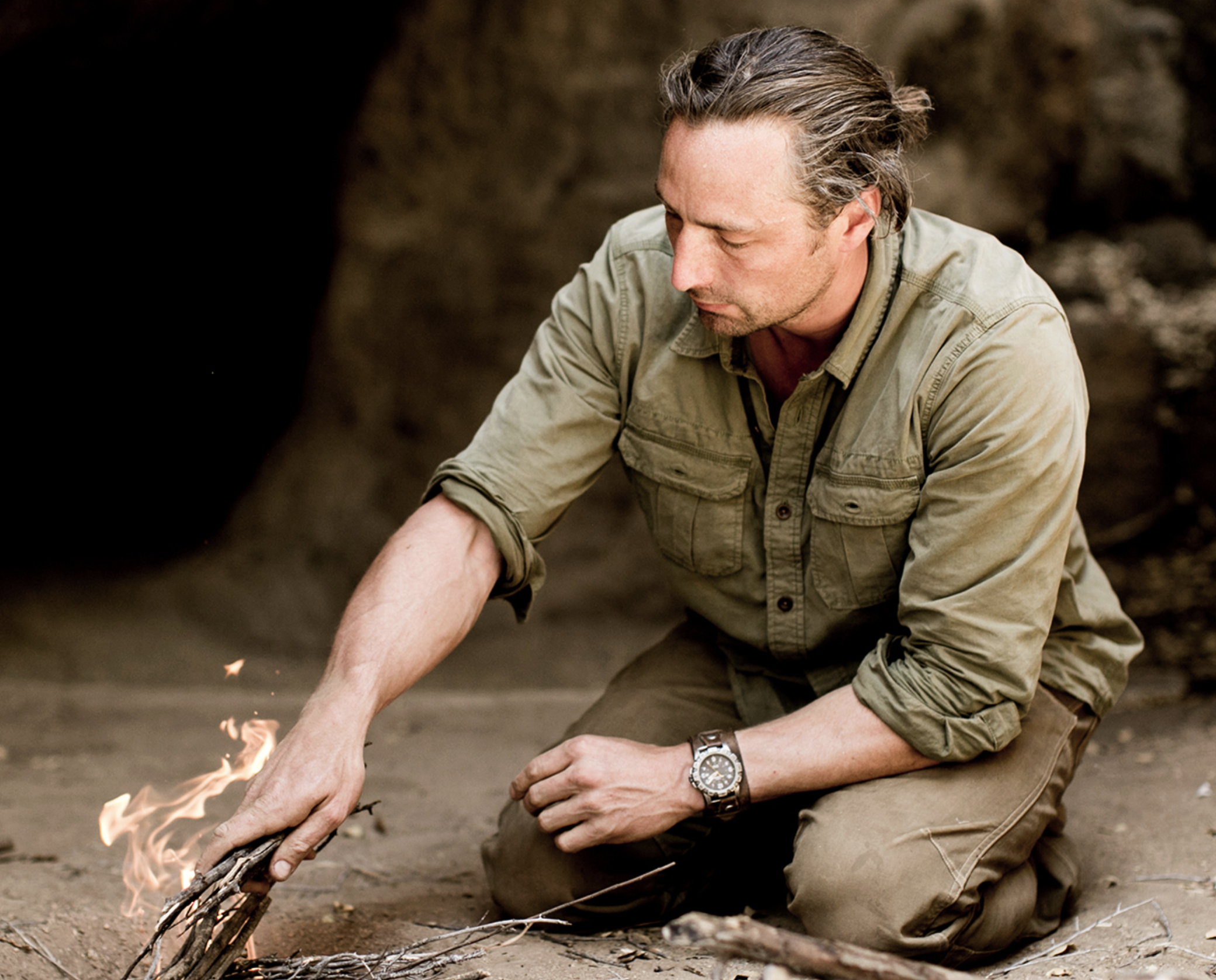 A man wearing a beige shirt and khaki pants kneeling outdoors, tending to a small fire with sticks in a natural setting with rocks in the background.