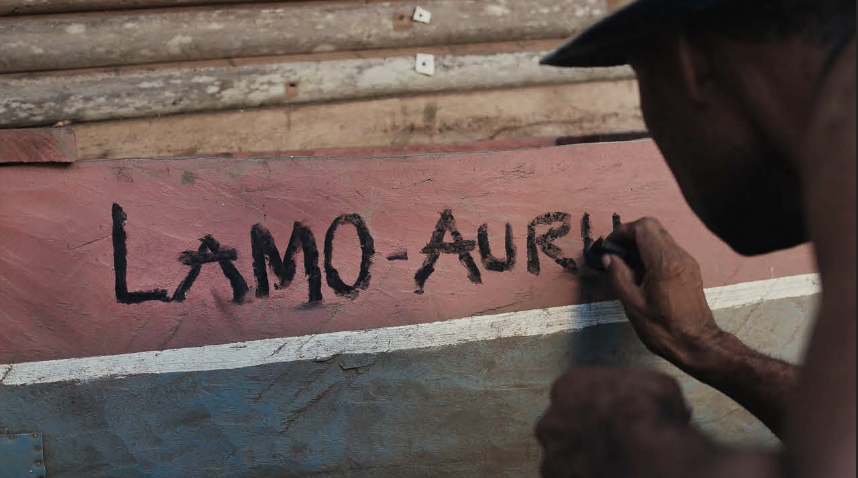 A man writing on a wooden boat with charcoal that says "LAMO-AURU".