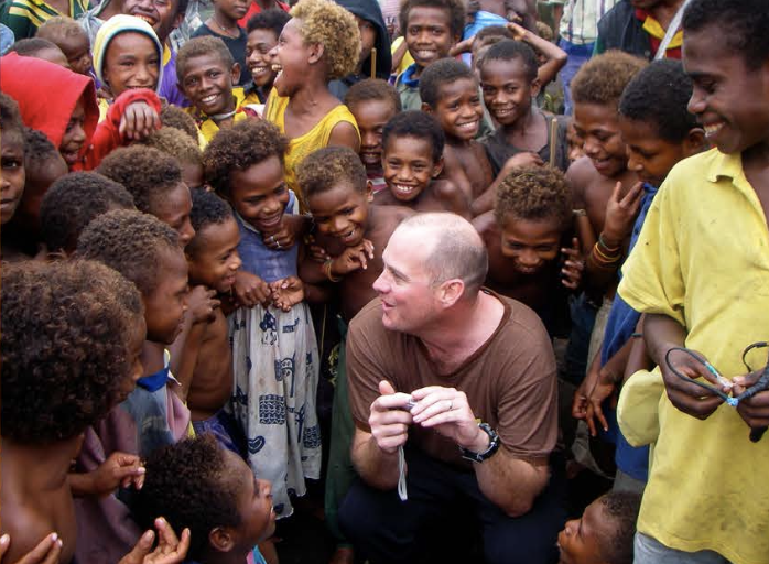 A man is surrounded by a group of smiling children in a tropical outdoor setting, engaging in conversation.
