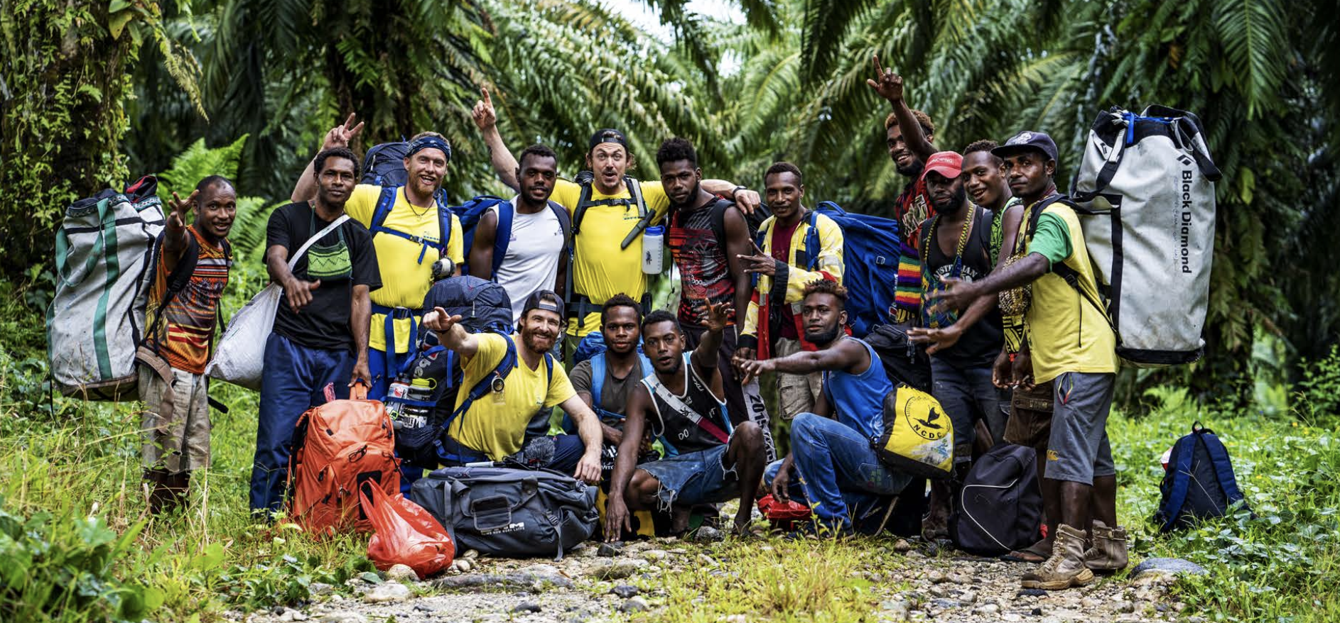 A diverse group of 16 people, including men and women of various ethnicities, posing together in a lush, green jungle of Lamo Auru. They wear outdoor gear with backpacks and hiking equipment, some making peace signs and smiling at the camera.