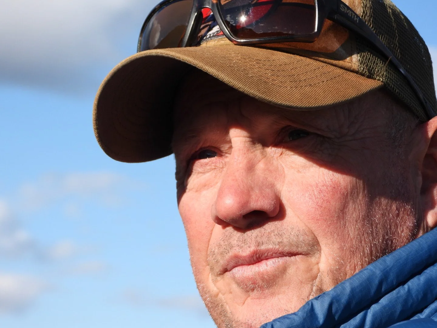 Close-up of a man wearing a tan baseball cap and sunglasses resting on the cap, with a blue sky in the background.