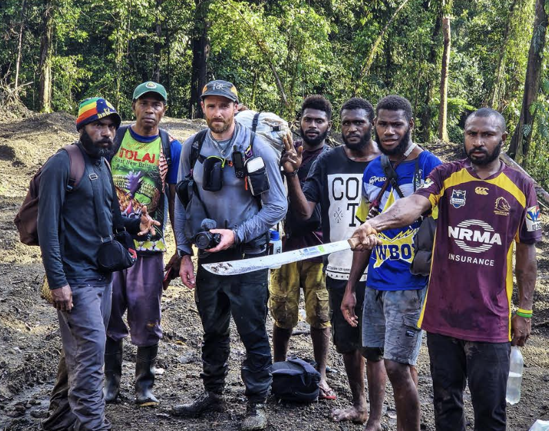 Group of eight men standing on an illegal logging road surrounded by trees, some holding backpacks and water bottles, one man is holding a machete, visibly upset by the destruction