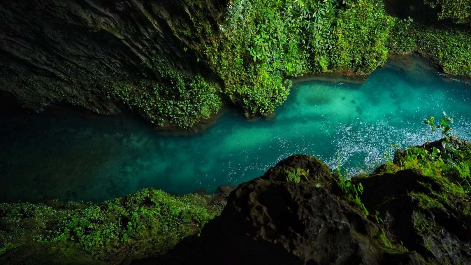 Aerial view of a turquoise river in the Lamo Auru Caldera from above surrounded by green moss and foliage on rocky cliffs.