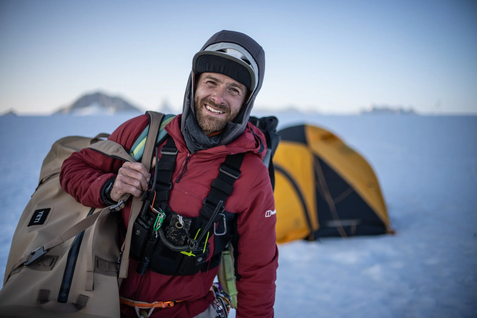 Smiling Waldo Etherington in red winter gear and a beanie outdoors in snow, carrying a backpack, with a yellow tent in the background.