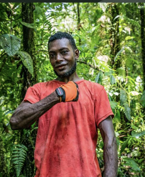 A man with dark skin, short curly hair, and a beard, wearing a red shirt and an orange glove, standing in a lush green jungle.
