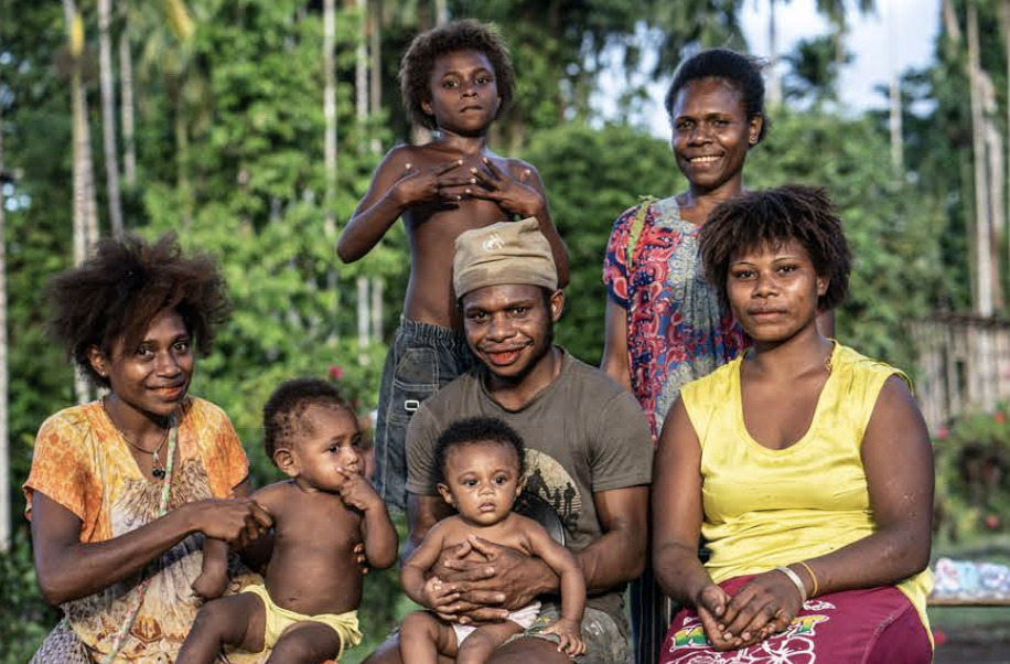 A group of people, including children and adults, outdoors in the lush, green Lamo Auru Forest with trees, smiling for a photo.