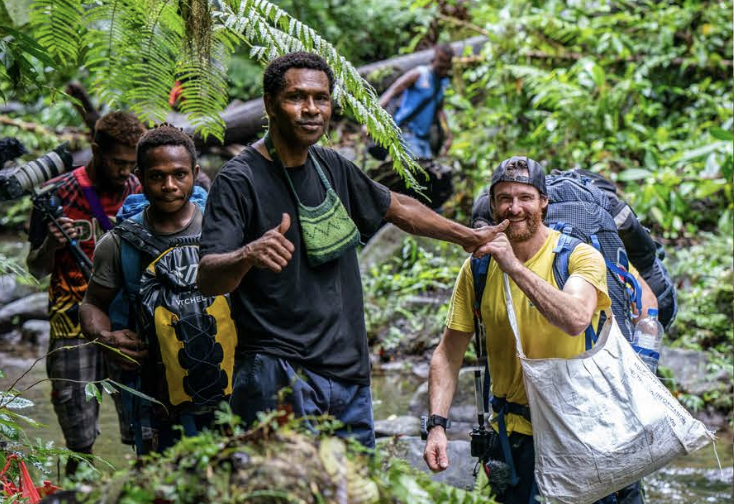 A group of hikers in a lush green rainforest, with two men in the front high-fiving each other. The man on the left wears a black shirt and the man on the right wears a yellow shirt and a cap, both carrying backpacks.