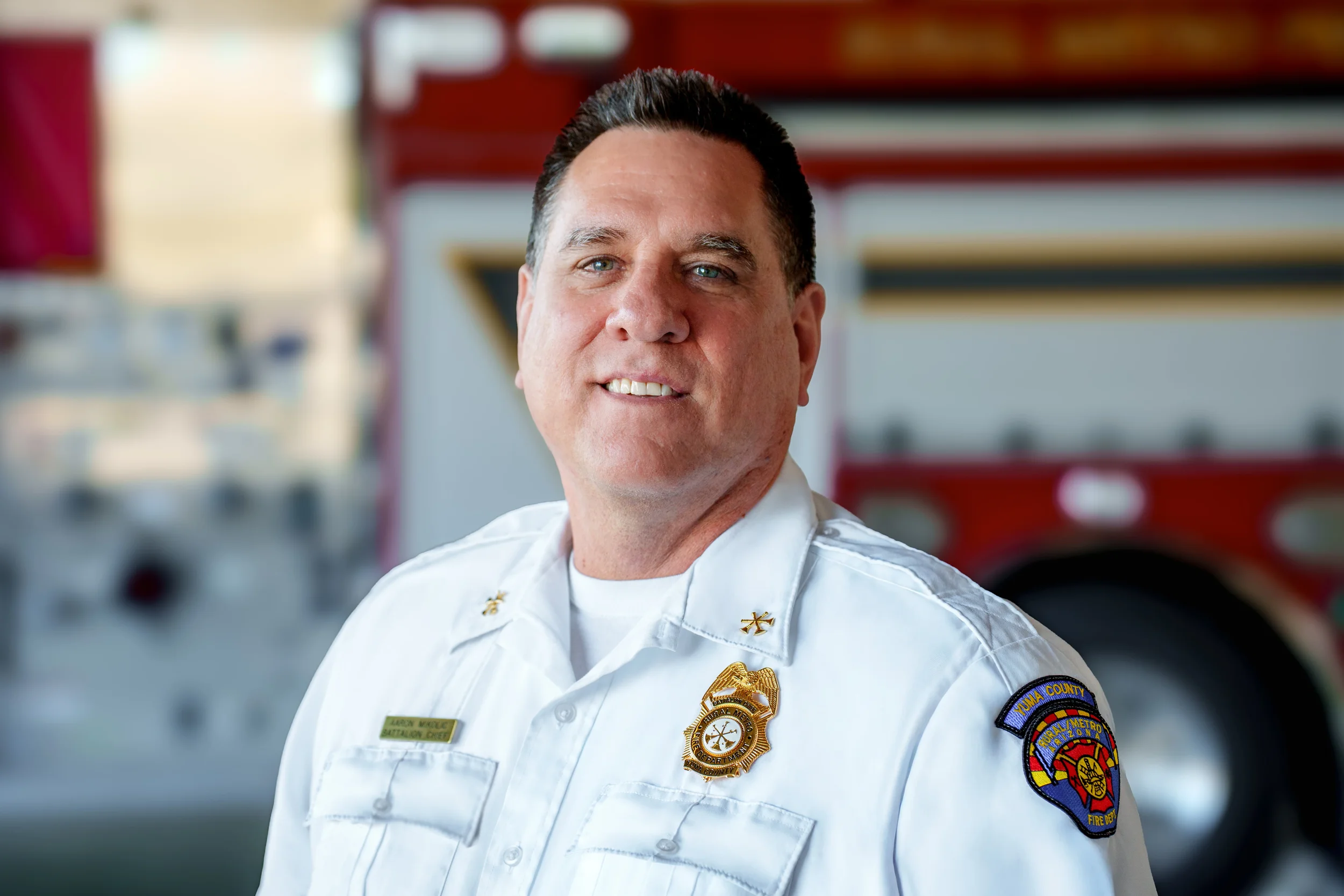 A smiling firefighter in uniform standing in front of a fire truck.