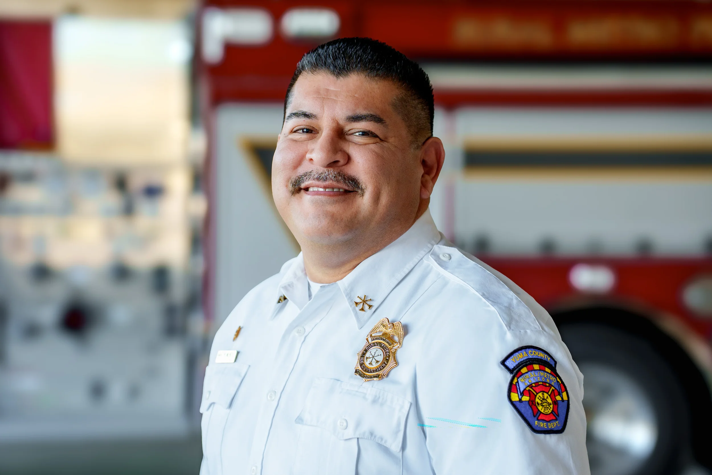 A smiling firefighter in uniform standing in front of a fire truck.