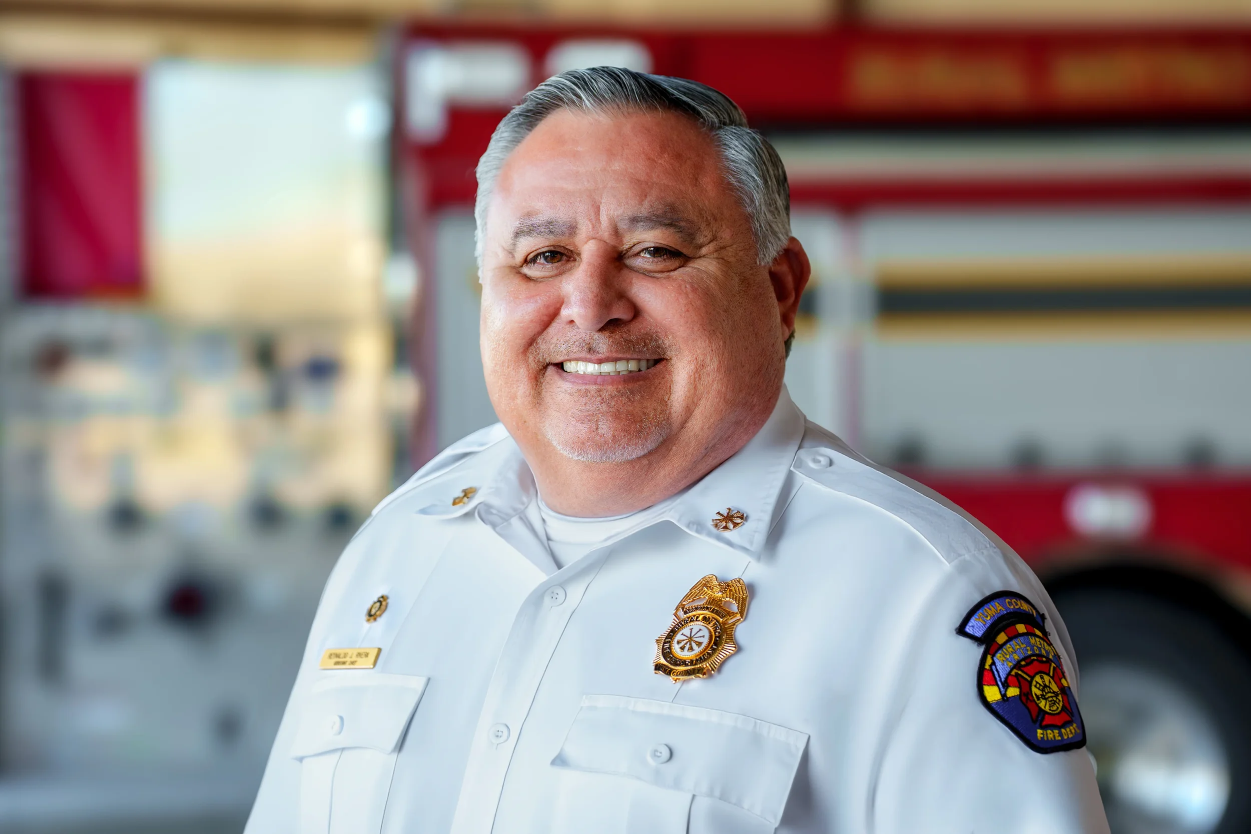 A smiling firefighter in uniform with a badge, standing in front of a red fire truck.