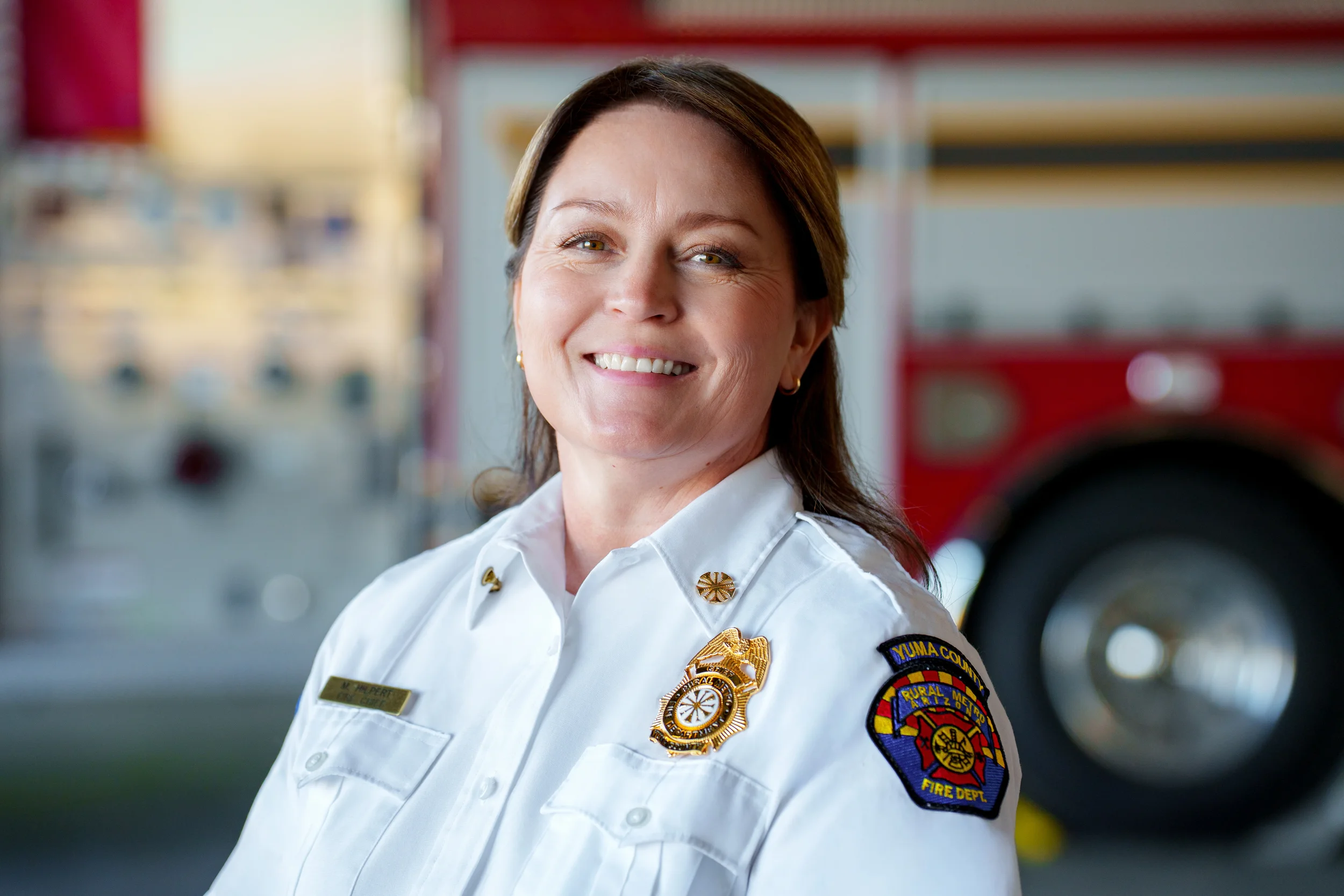 A smiling female firefighter in uniform with a fire truck in the background.