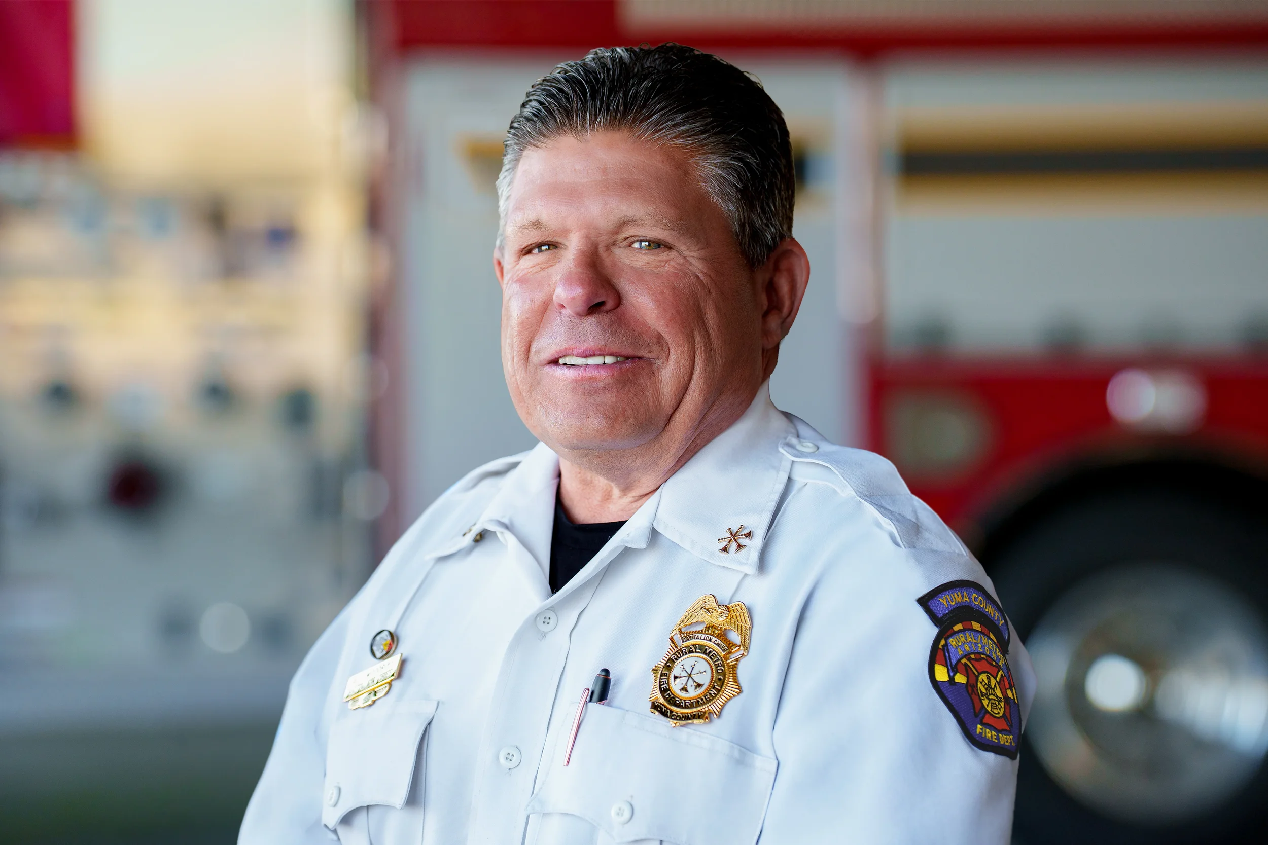 A firefighter in uniform standing in front of a fire truck, smiling at the camera.