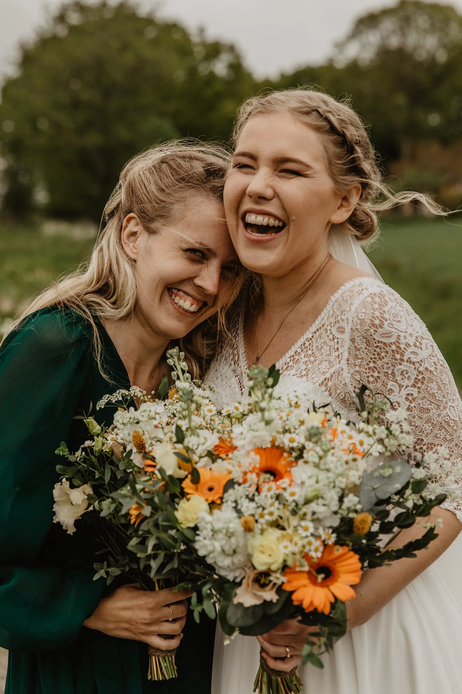 Två kvinnor, en i en brudklänning och en i en mörk grön klänning, leker gladeligen tillsammans medan de håller blomsterbuketter, utomhus.
