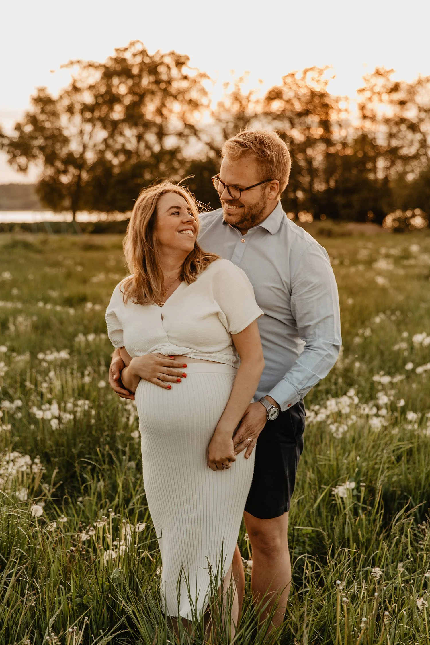 En gravid kvinna och en man som håller om henne, står i en blommande gräsmark under solnedgången, ler mot varandra.