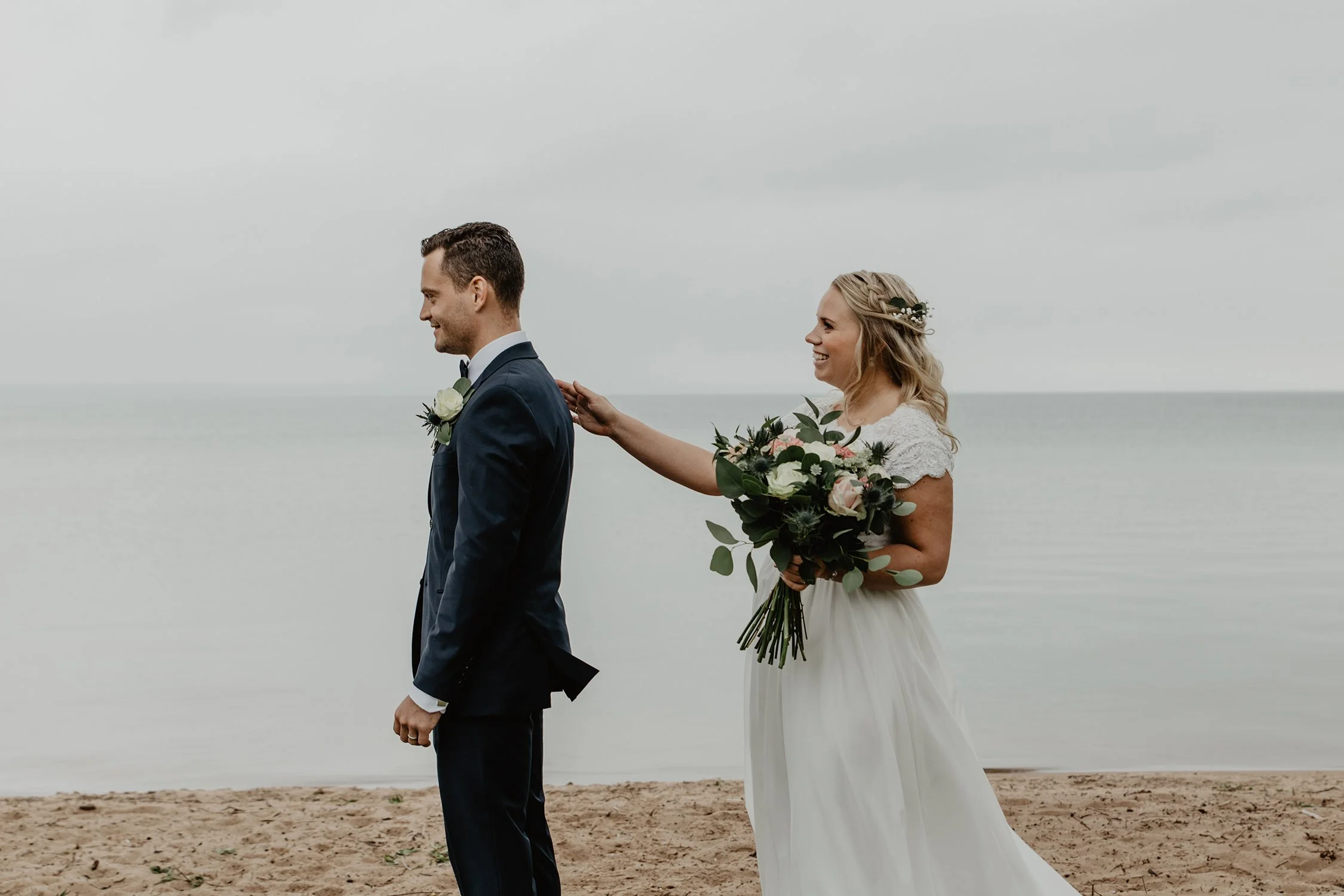 En brud och en brudgummi står på Vätterstranden under en First look, bruden håller sin hand på brudgummins axel och han ska snart vända sig om.