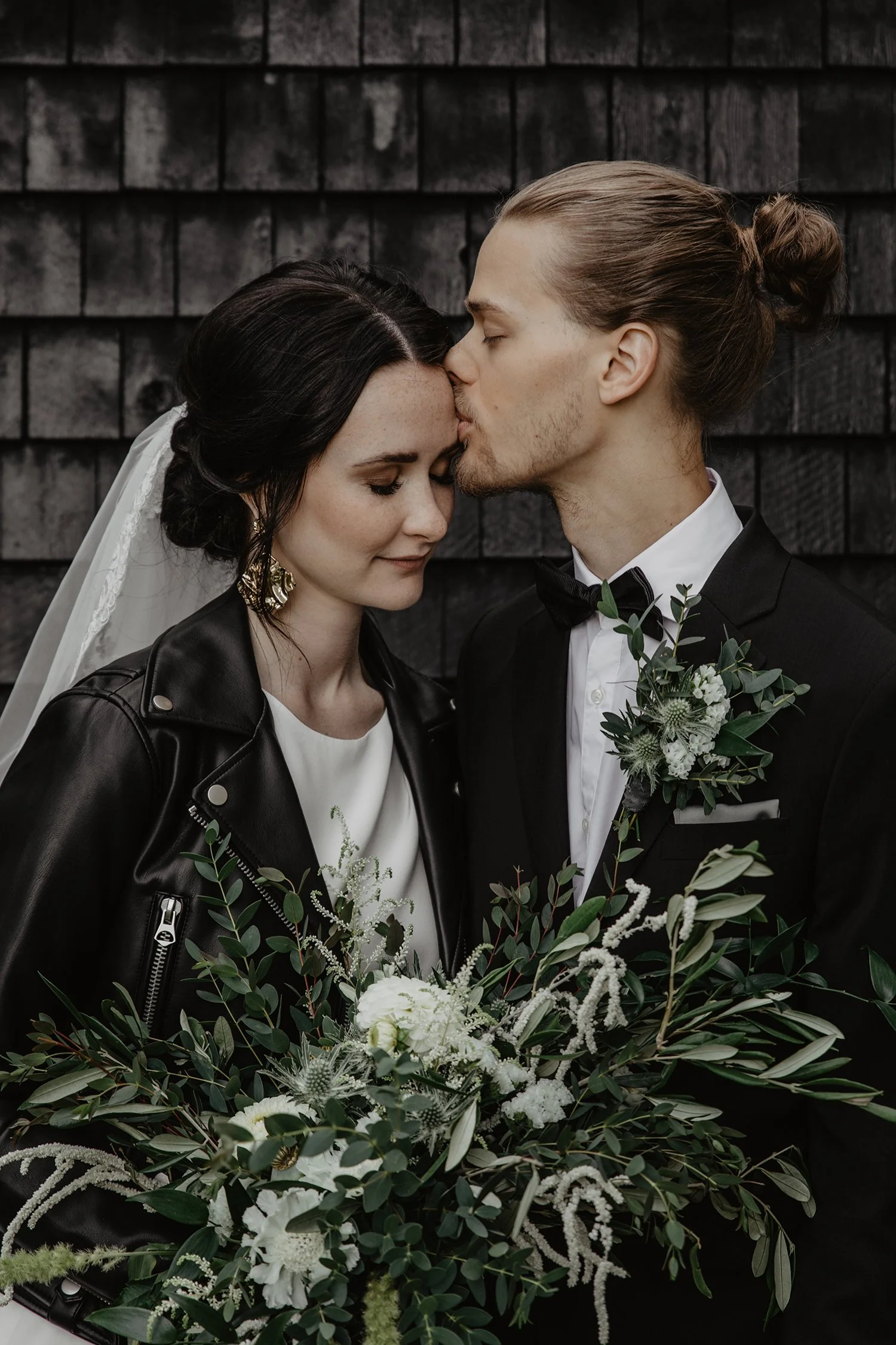 Brudparet står nära varandra och brudgummen kysser brudens panna. Bakom är en trävägg i svart och bruden har en svart skinnjacka på sig. Fotograferingen ägde rum i Sävsjö vid en kyrka.