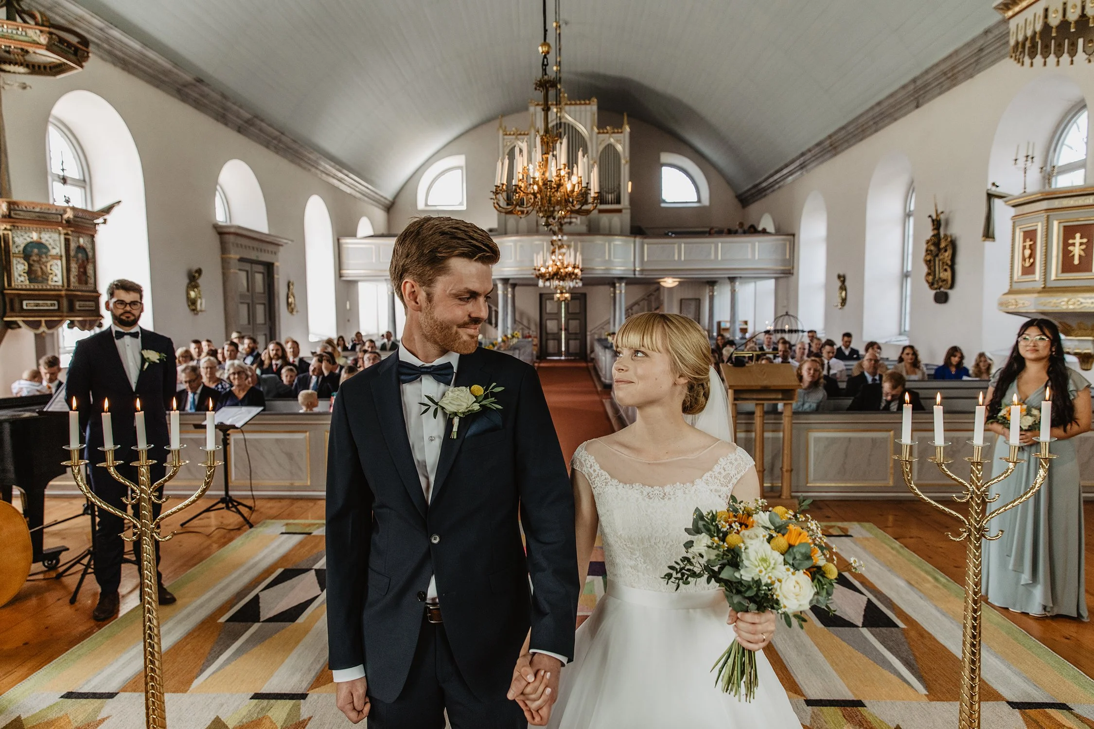 En nygift brud och brudgum håller handen under ett bröllopsceremoni i en kyrka med många åskådare i bakgrunden.