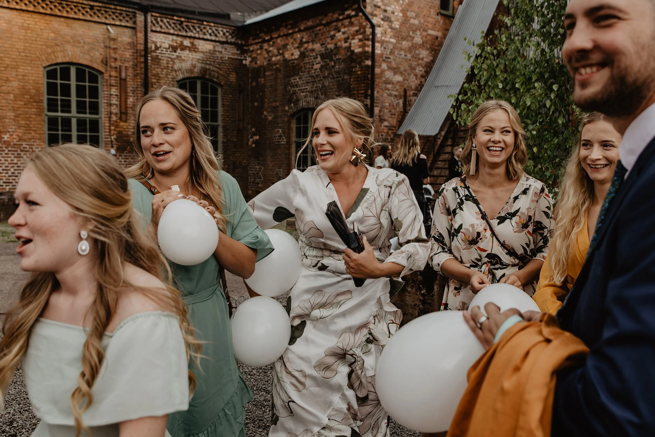 Grupp människor väntar på brudparet utanför bröllopslokalen. De är glada och skrattar, håller vita ballonger med Åminne Bruk bakom sig på en bröllopsfotografering i Värnamo.