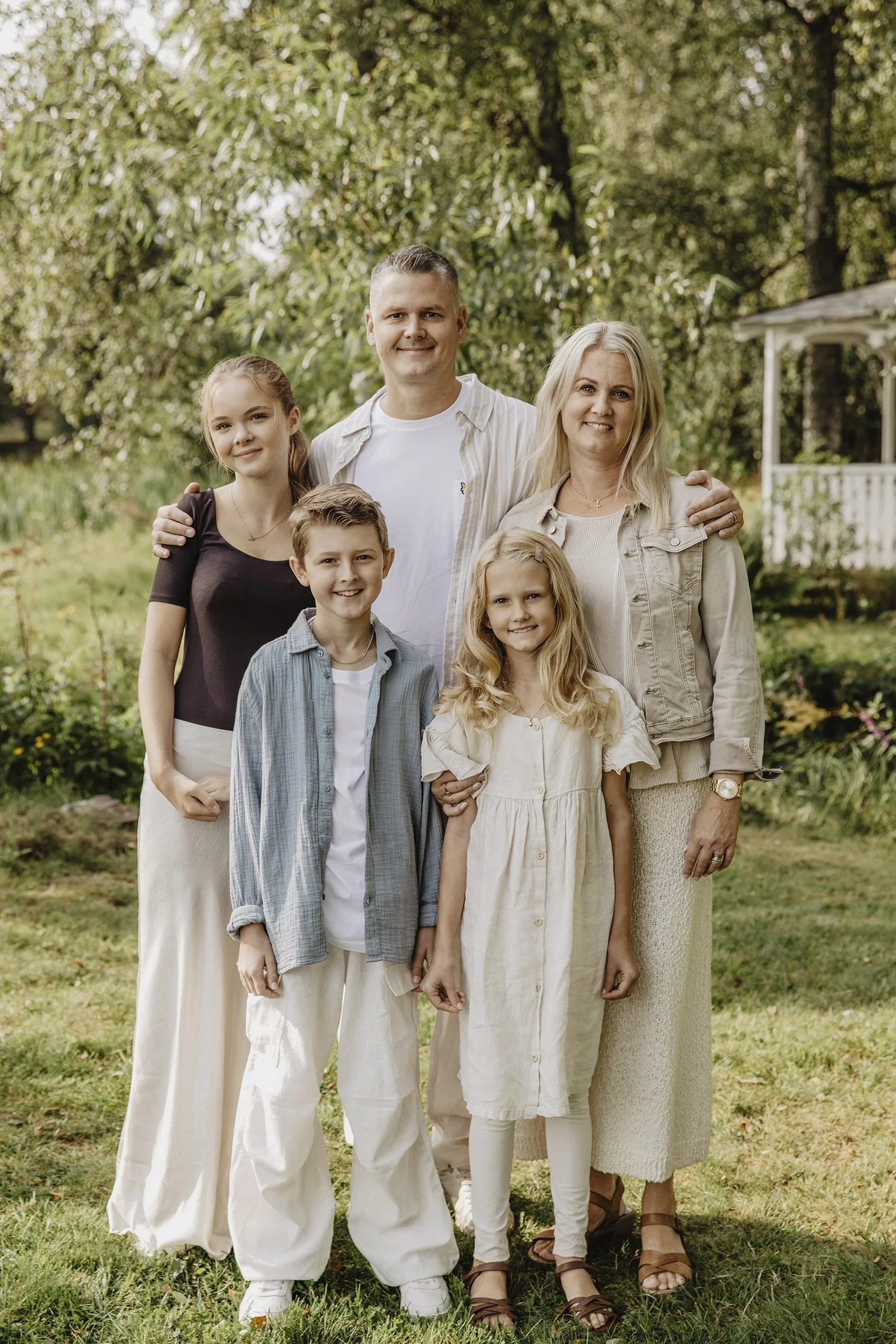 En familj med två vuxna och tre barn står utomhus i en trädgård, ler och tittar mot kameran.