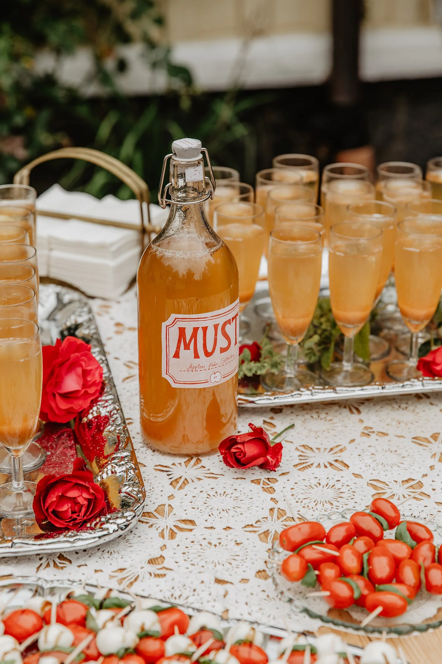En glasflaska med äppelcider eller äppelvin med ett etikett som säger 'MUST' står framför flera glas fyllda med en liknande dryck. Det är dekorerat med röda rosor på en vit, spetsad duk. Det finns även en skål med små, röda och vita äppliga godisbita