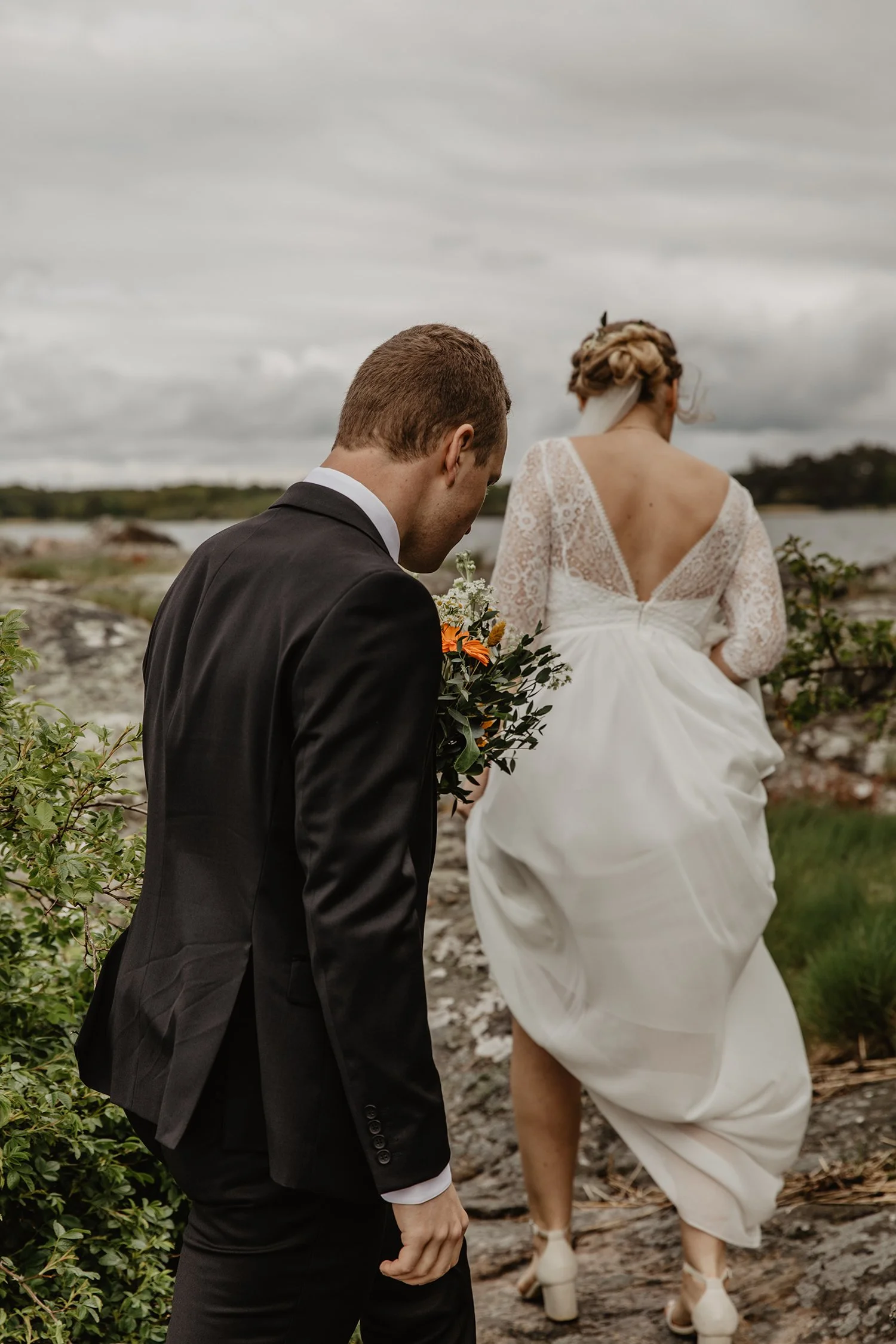En man i svart kostym håller ett blomsterarrangemang medan en kvinna i en vit brudklänning går bortanför honom på en stenig strand med ett vatten och molnig himmel i bakgrunden.