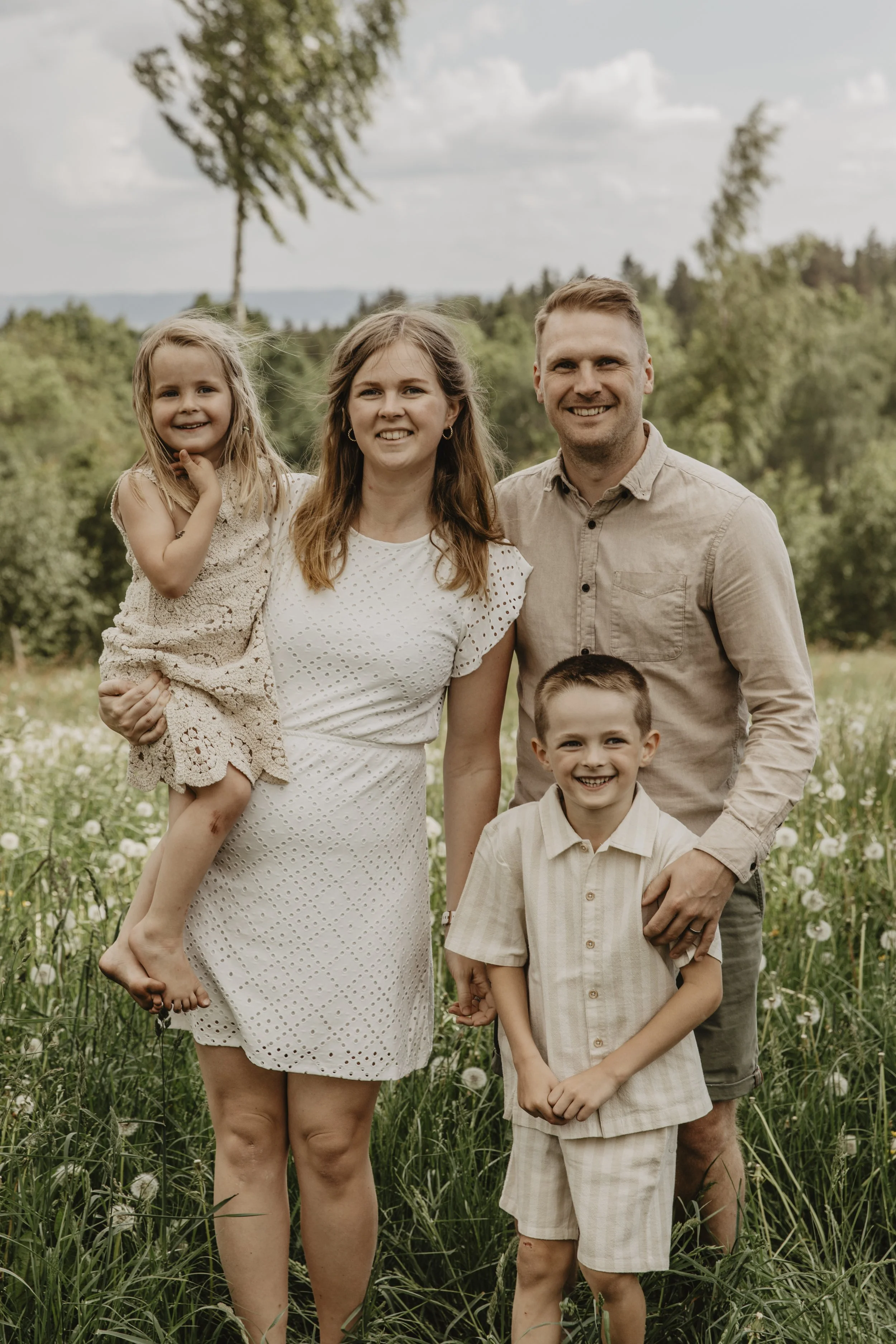 En familj på fem som står i ett grönt fält med träd i bakgrunden, alla ler och poserar för kameran.